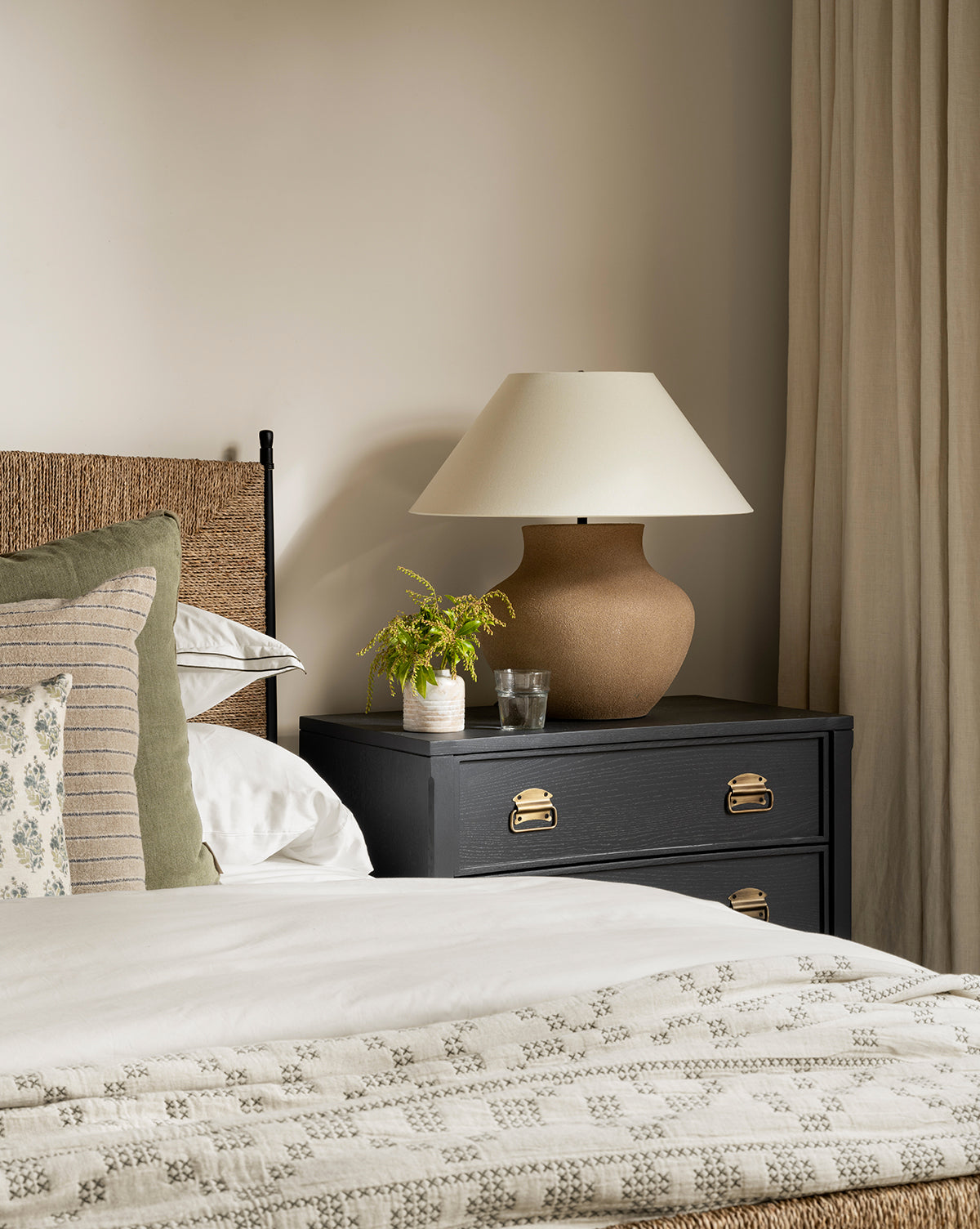 A cozy bedroom corner features a bed with patterned pillows, a black nightstand holding the Parma Table Lamp (textured ceramic base, ivory linen shade), a small plant, and water glasses set against cream walls and beige curtains, by McGee & Co.