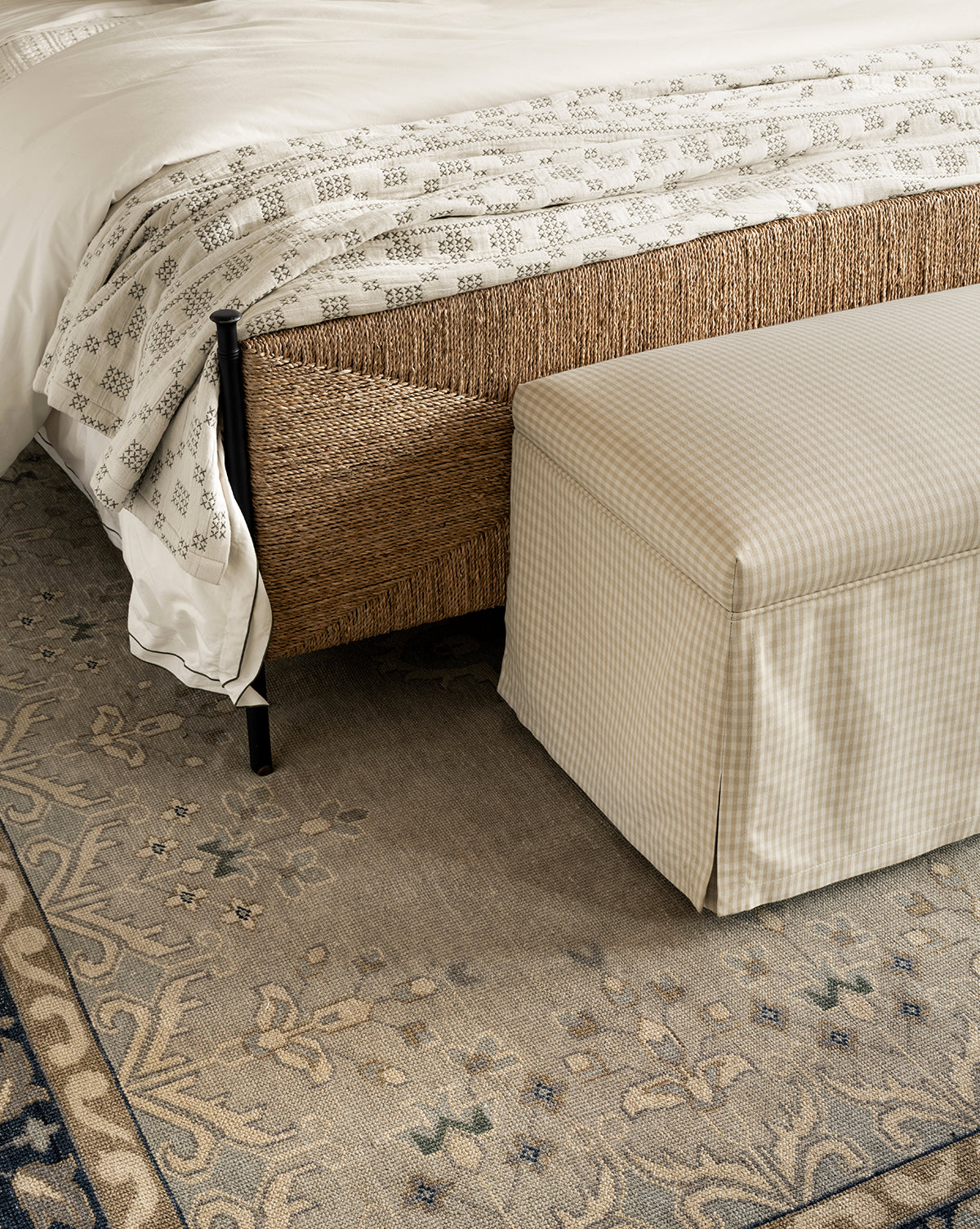 A beige checkered bench sits at the foot of a bed with a woven frame and white patterned bedding, all atop the Deveraux Hand-Knotted Wool Rug in neutral tones, by McGee & Co.