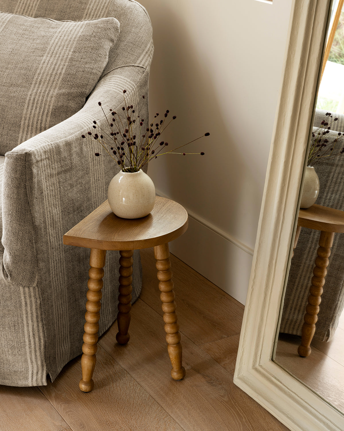 The Makerspalm Alice Honey Wood Stool, a compact ash wood accent with three spindle legs, displays a round beige vase with dried branches beside a gray-striped armchair and large framed mirror on a wooden floor, McGee & Co.