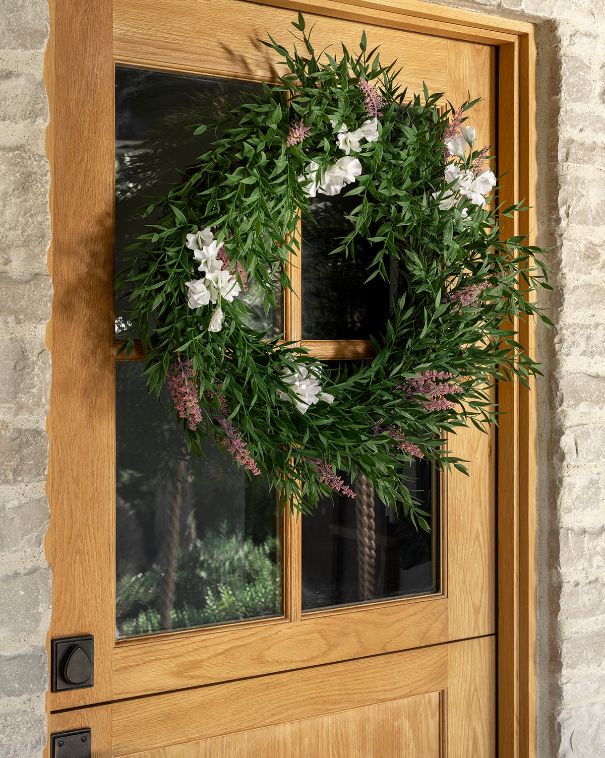 The Faux Bamboo & Sweet Pea Wreath, adorned with green leaves and sweet pea flowers, hangs on a glass-paneled wooden door set in a stone exterior wall, by McGee & Co.