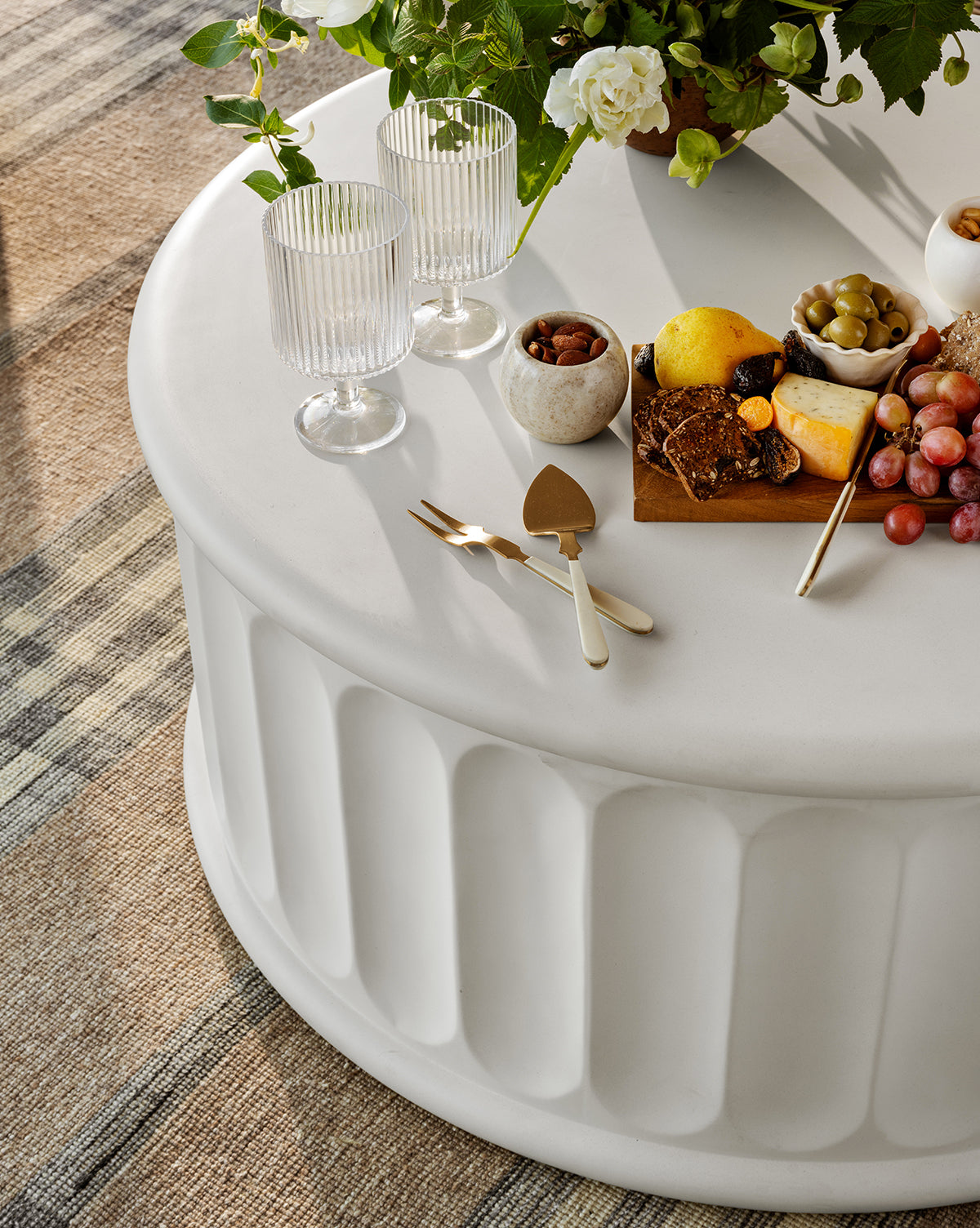 A round white table set with two Sanibel Acrylic Goblets, a small bowl of nuts, gold cutlery, a wooden board holding cheese, crackers, grapes, lemon, and olives, with a floral arrangement in the background.