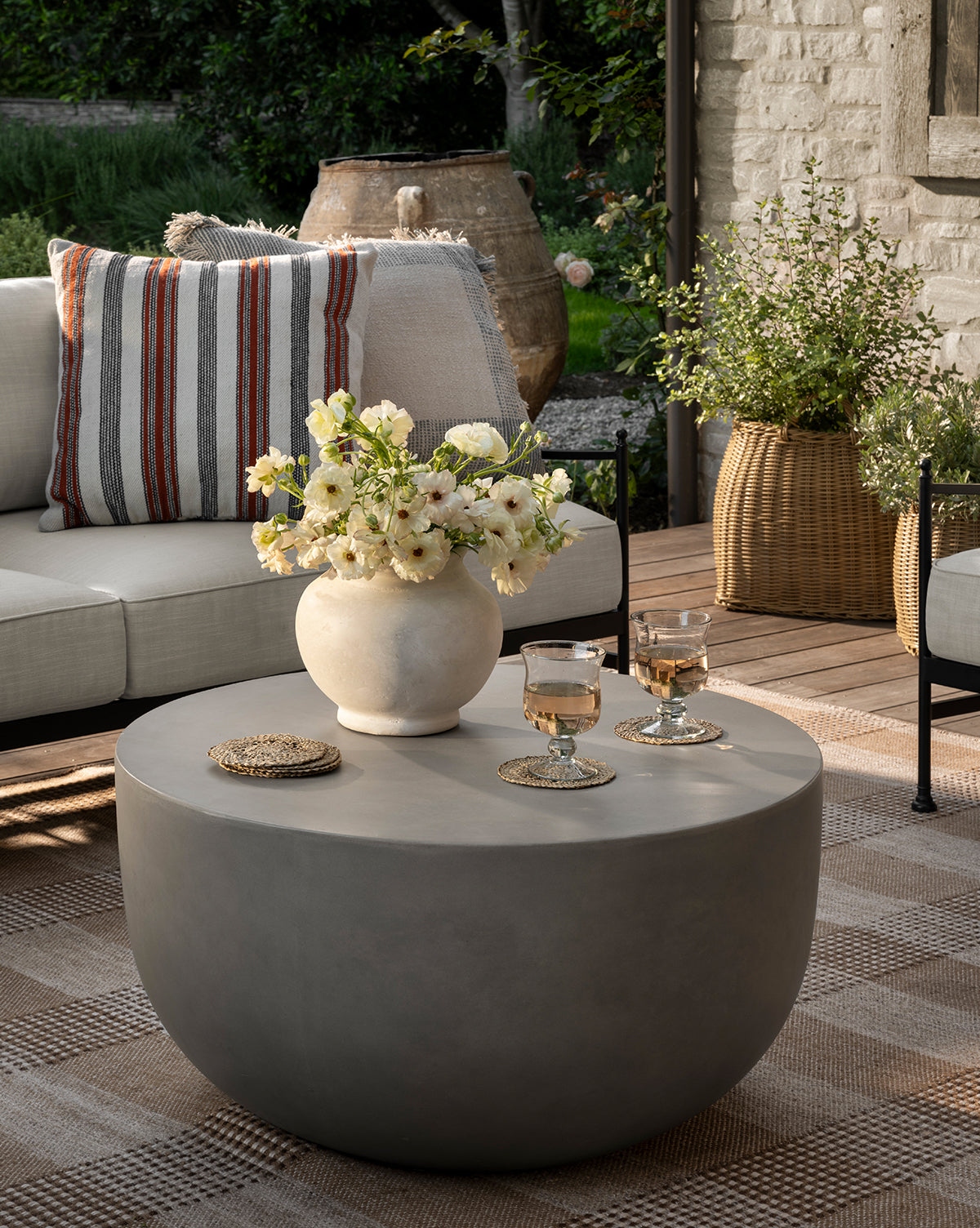 A modern patio showcases a round gray coffee table with a Nola Paper Mache Vase holding white flowers and two glasses, while a beige sofa with striped pillows and lush potted plants create a cozy, inviting outdoor space, by McGee & Co.