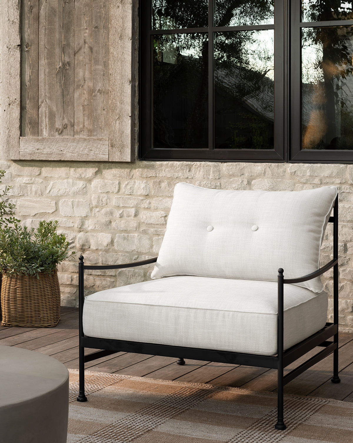 The Pomeroy Outdoor Lounge Chair with white performance cushions and a black powder-coated steel frame sits on a checkered rug by a basketed plant, in front of a stone wall and large window with wooden shutter by McGee & Co.