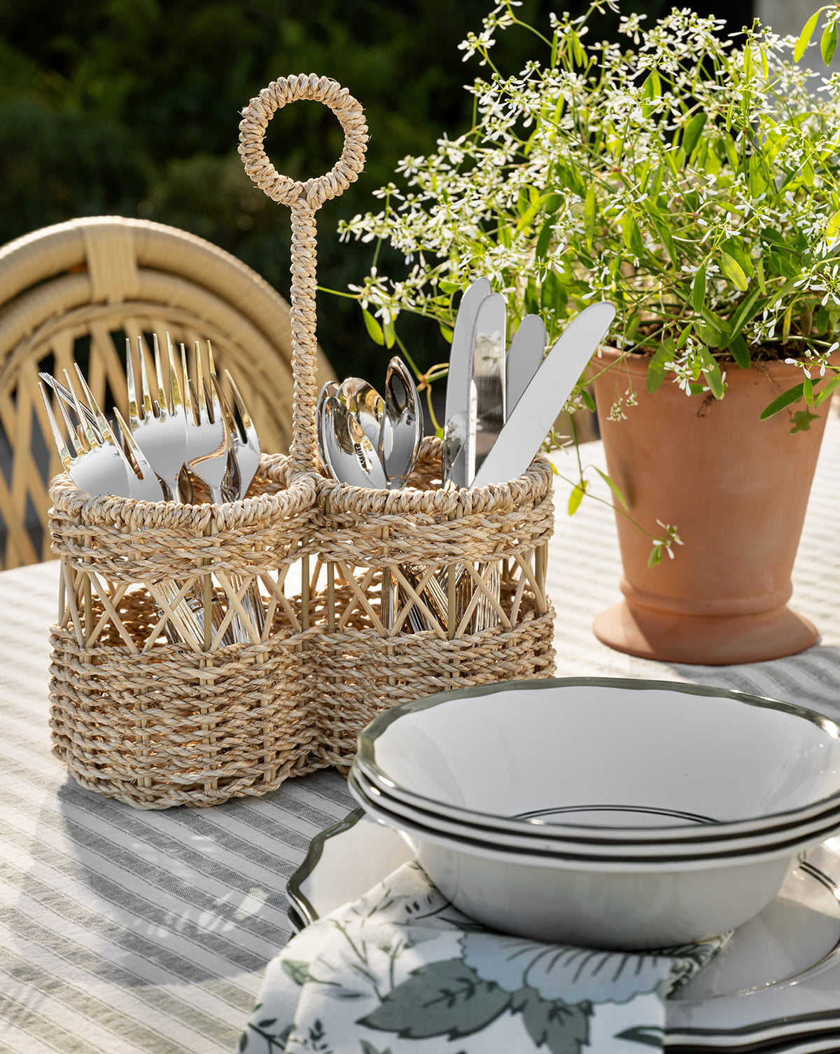 The Stokes Woven Caddy with silverware sits on a striped tablecloth beside stacked white plates and a potted plant, creating an inviting outdoor dining setting, by McGee & Co.