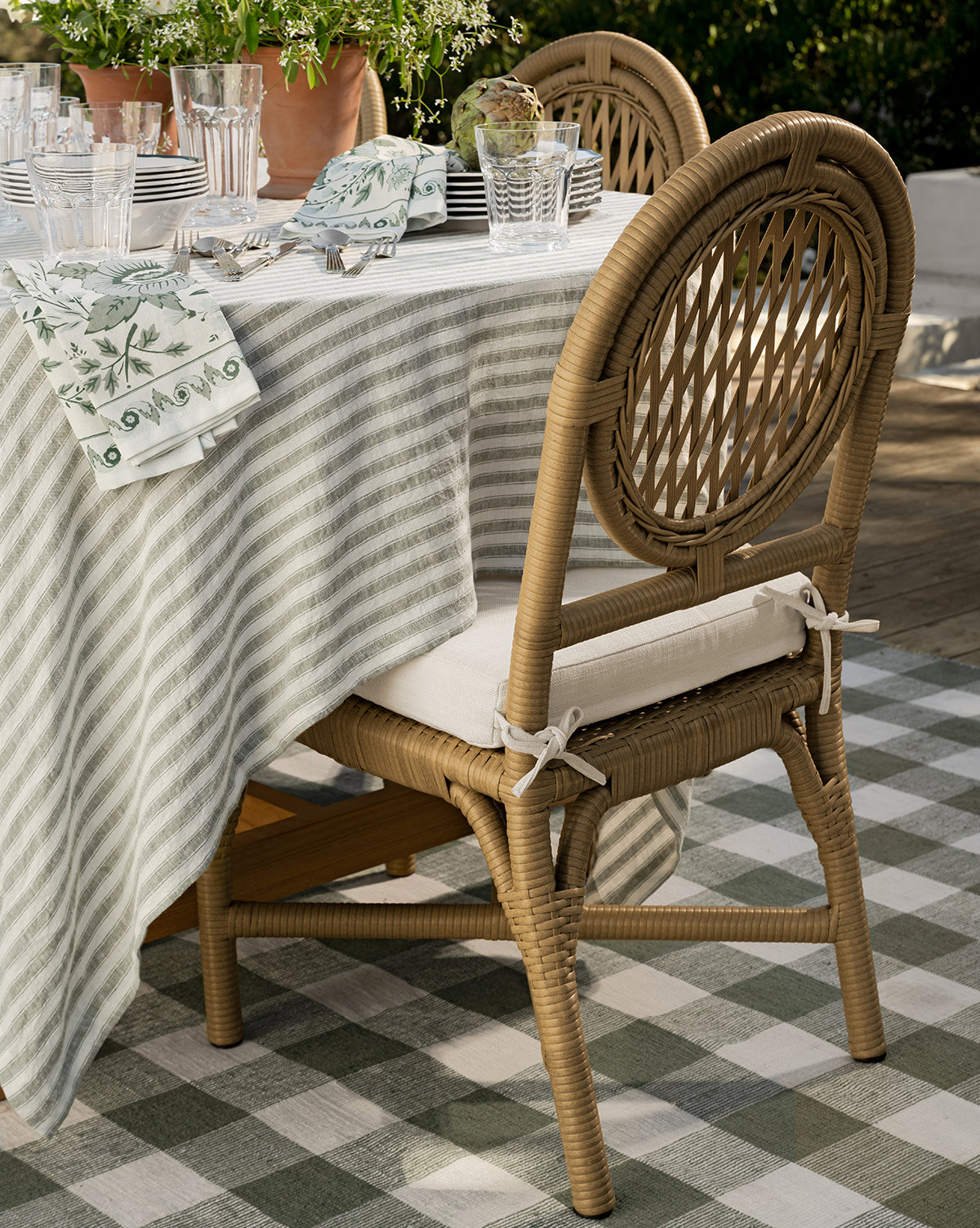 Two Marisol Indoor/Outdoor Dining Chairs with cushions sit at a table set with a striped tablecloth, glassware, plates, and patterned napkins on a checkered rug surrounded by greenery, by McGee & Co. Weather-resistant and handwoven wicker design.