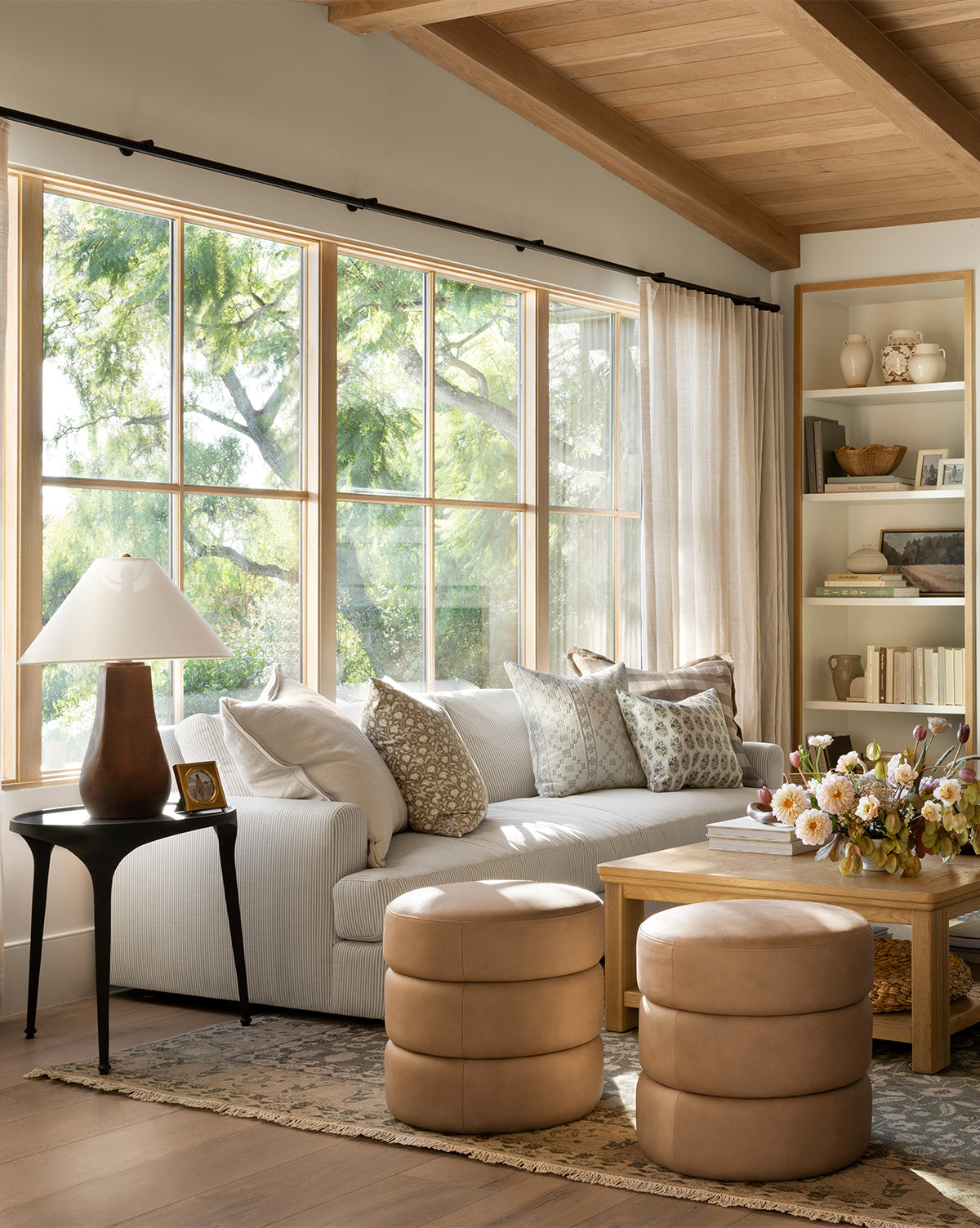 A bright living room with a large window, white sofa with patterned pillows, two Channel Ottomans, a wooden coffee table with a floral arrangement, bookshelves, and a side table topped by a lamp by McGee & Co.