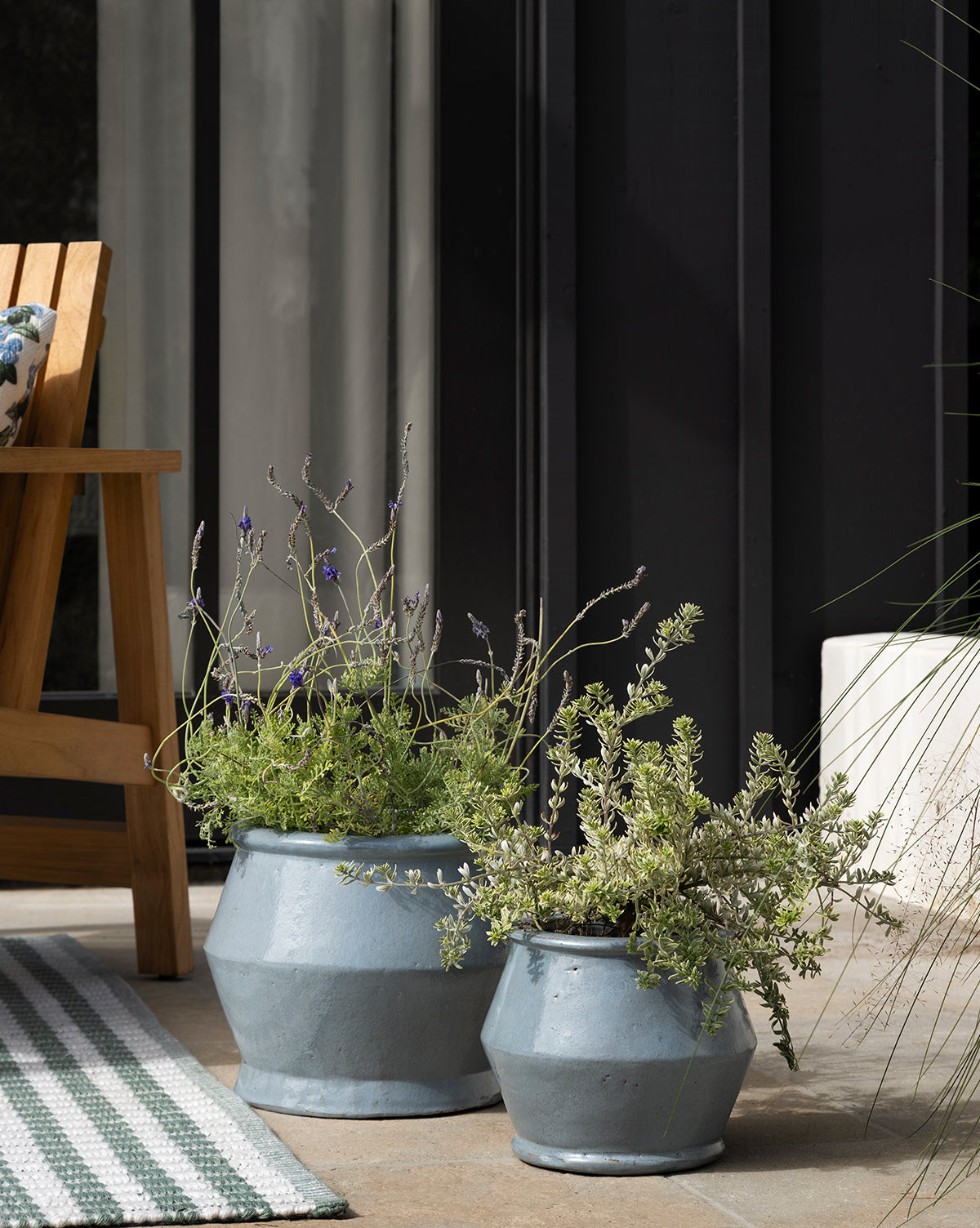 Two UNITED POTTERIES SAIGON Corsica Pots with green and lavender plants sit on a patio beside a wooden chair and striped rug, with glass doors framed in black metal in the background. McGee & Co.