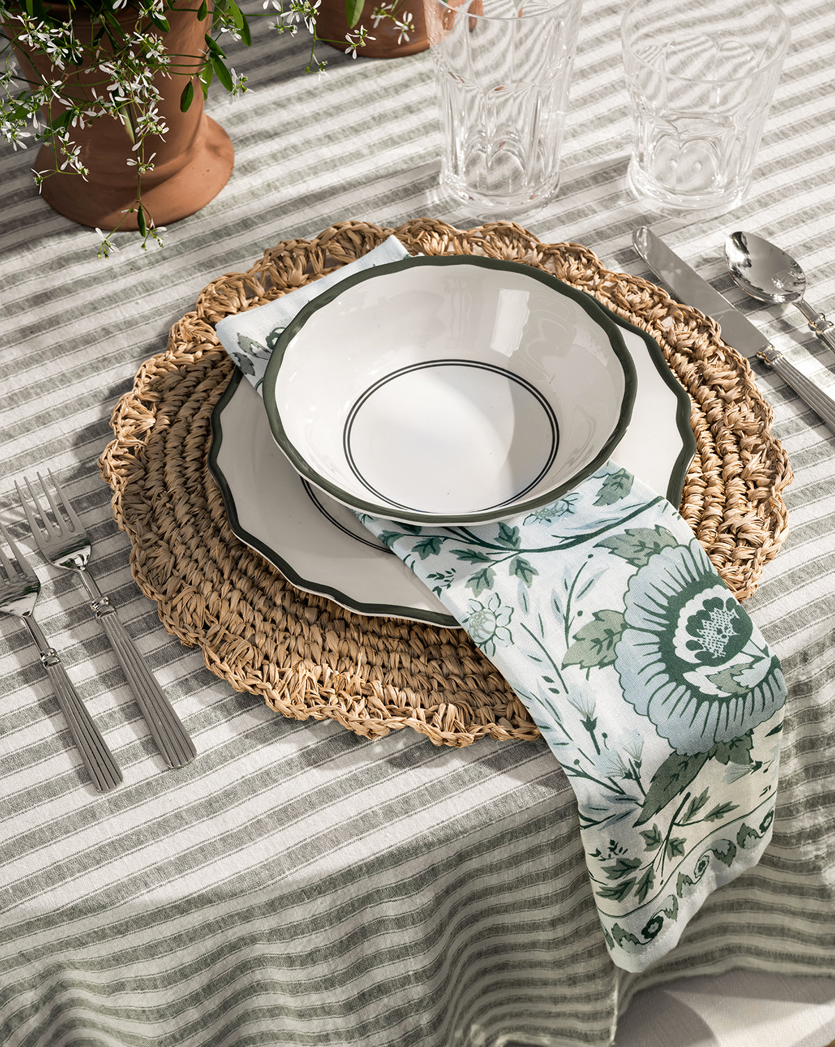 A table setting with a woven placemat, scalloped edge white plate, Alcott Melamine Bowls (Set of 4) with green rims, floral napkin, striped tablecloth, silverware, clear glasses, and potted plants in the background by McGee & Co.
