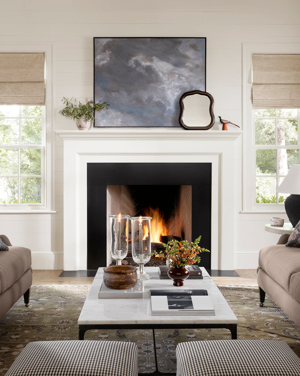 A cozy living room features a lit fireplace with two windows in beige shades. On the white mantel, the Freya Shelf Mirror is displayed alongside a landscape painting and plant. A coffee table with candles and books sits between two sofas, by McGee & Co.