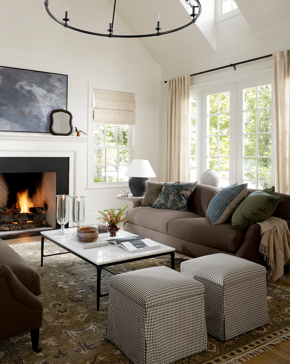 A cozy living room with a lit fireplace, brown sofas accented by green and blue pillows, a white marble coffee table, two Latimer Ottomans (Ready to Ship), a large rug, and beige curtains framing sunlit windows—ideal for home styling enthusiasts, by McGee & Co.