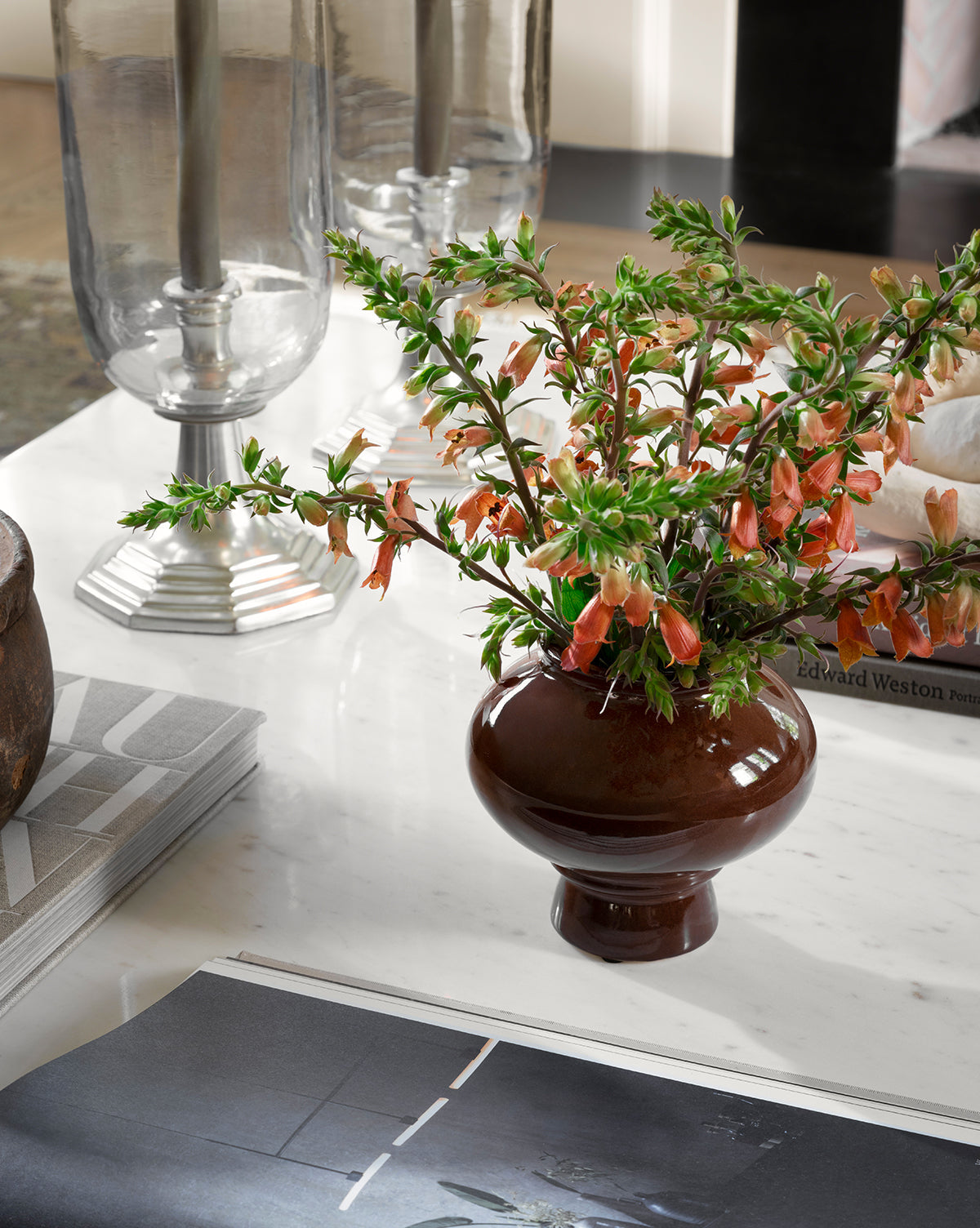 A Louis Vase with orange and green flowers sits on a white marble table beside magazines and silver candleholders, while an open book in the foreground adds elegance to this modern home decor scene, by McGee & Co.