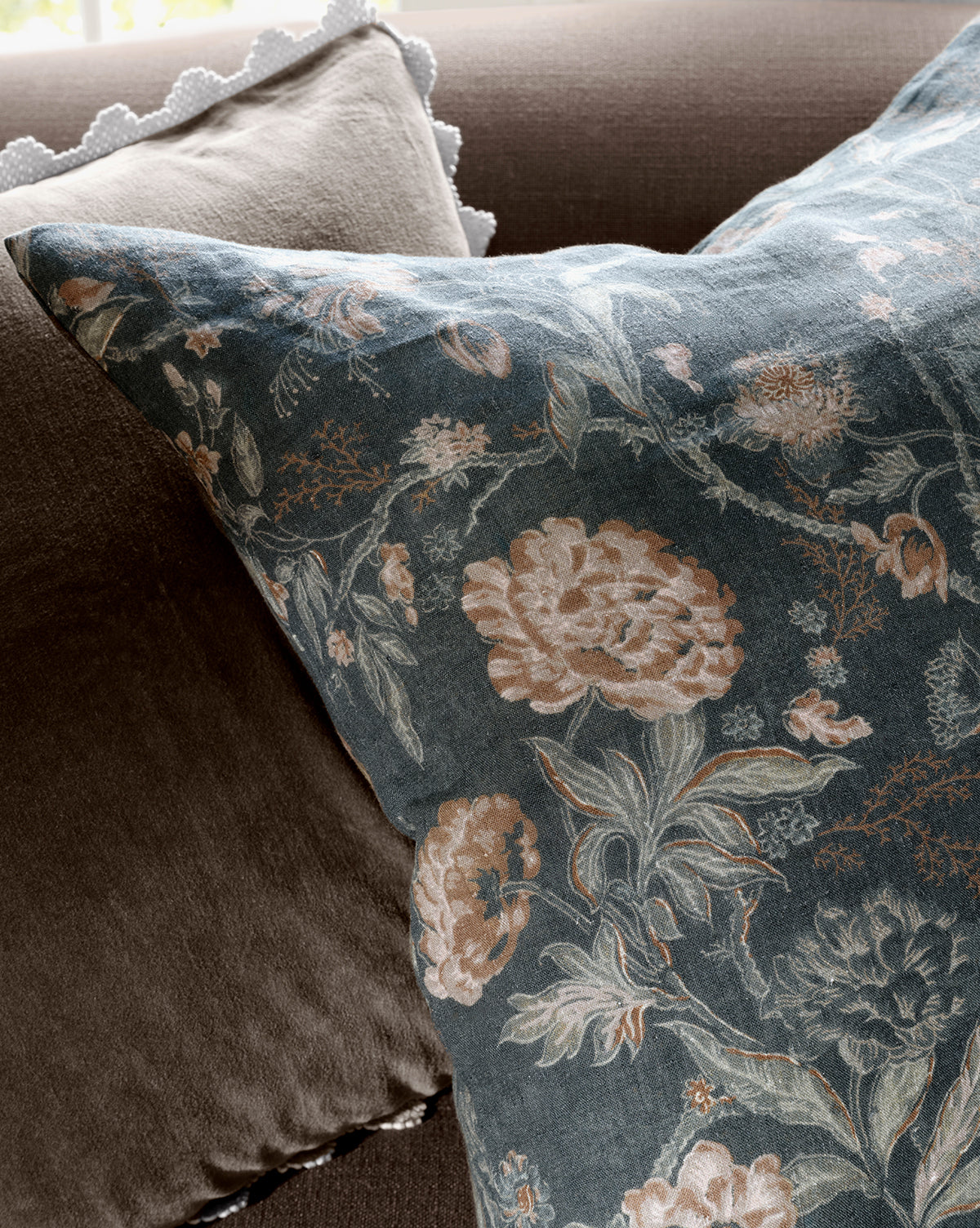 A close-up of the Hestercombe Pillow Cover, showcasing vintage-inspired muted pink and beige flowers on a dark background, paired with a plain brown pillow and a white scalloped-edged cushion in the background, by McGee & Co.