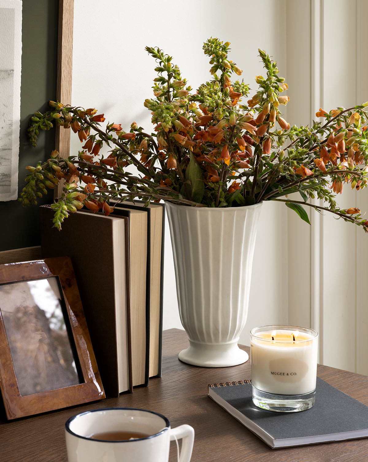 The Delphi Footed Vase, crafted from white stoneware and filled with orange flowers, rests on a wooden table beside a candle, closed notebook, mug, framed photo, and upright books by McGee & Co.