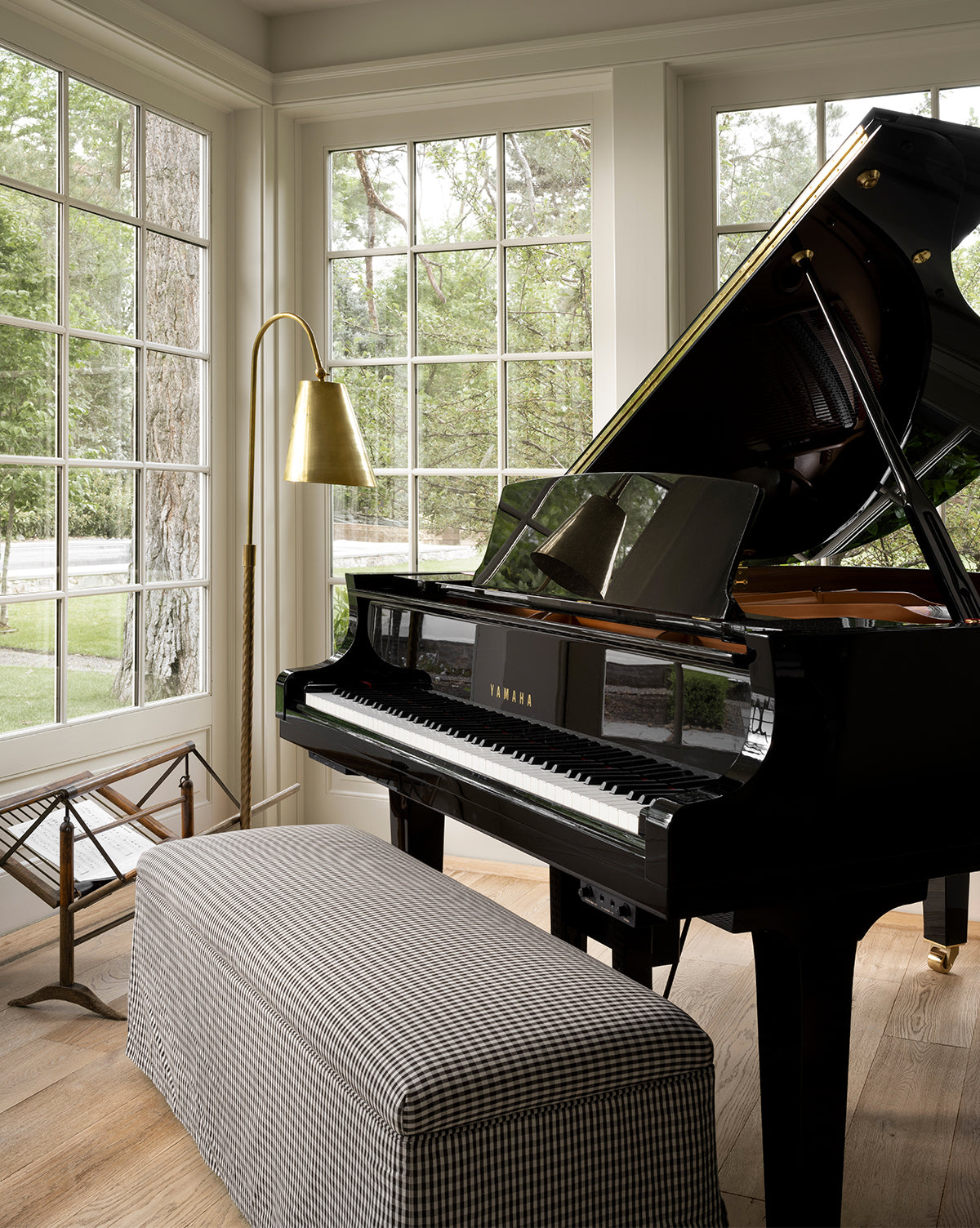 A black Yamaha grand piano sits by large windows in a bright room, next to the Saylor Floor Lamp and a padded bench with a checkered fabric cover; greenery is visible outside, by McGee & Co.