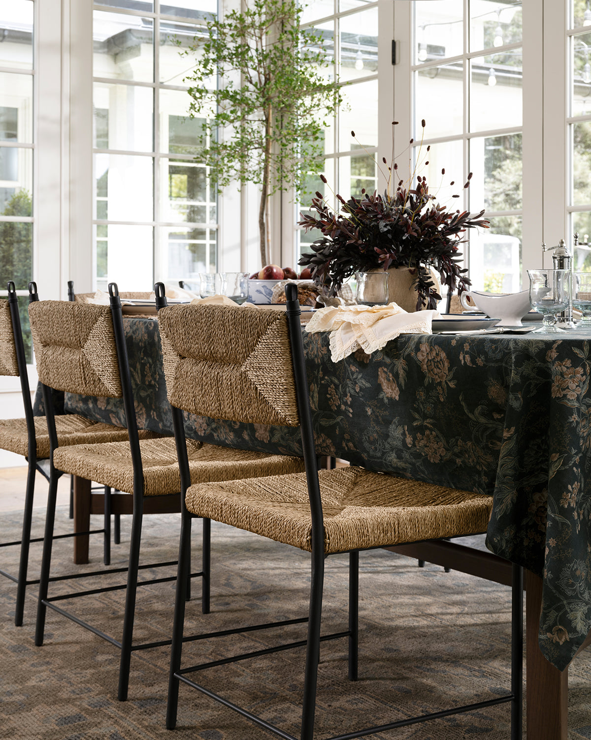 A dining table set with a dark floral tablecloth, Peregrine Chairs, glassware, plates, and a centerpiece of dark foliage in a bright room with large windows and a potted tree by McGee & Co.