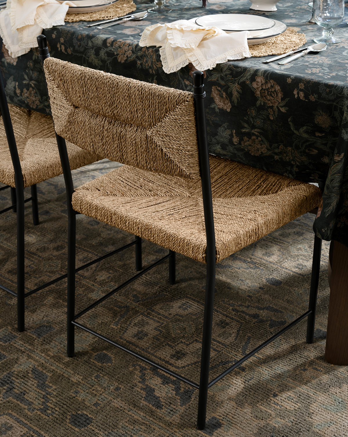 The Peregrine Chair, crafted from woven seagrass with a black metal frame, sits at a dining table adorned with a dark floral tablecloth, plates, silverware, and napkins. A patterned rug adds texture to the floor beneath, by McGee & Co.