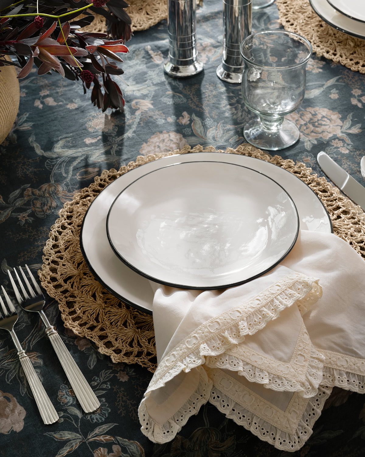 A place setting on a floral tablecloth features a white plate on a woven placemat, silver cutlery, a clear water glass, Lace Trim Napkins (Set of 4), and a vase with dark foliage in the corner, by McGee & Co.