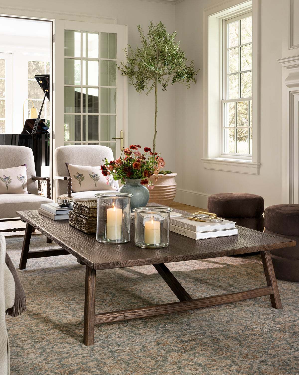 A cozy living room features a wooden coffee table with candles, books, and flowers. Neutral sofas, poufs, a potted tree, and the Endicott Hand-Tufted Wool Rug enhance the bright space filled with natural light from large windows by McGee & Co.