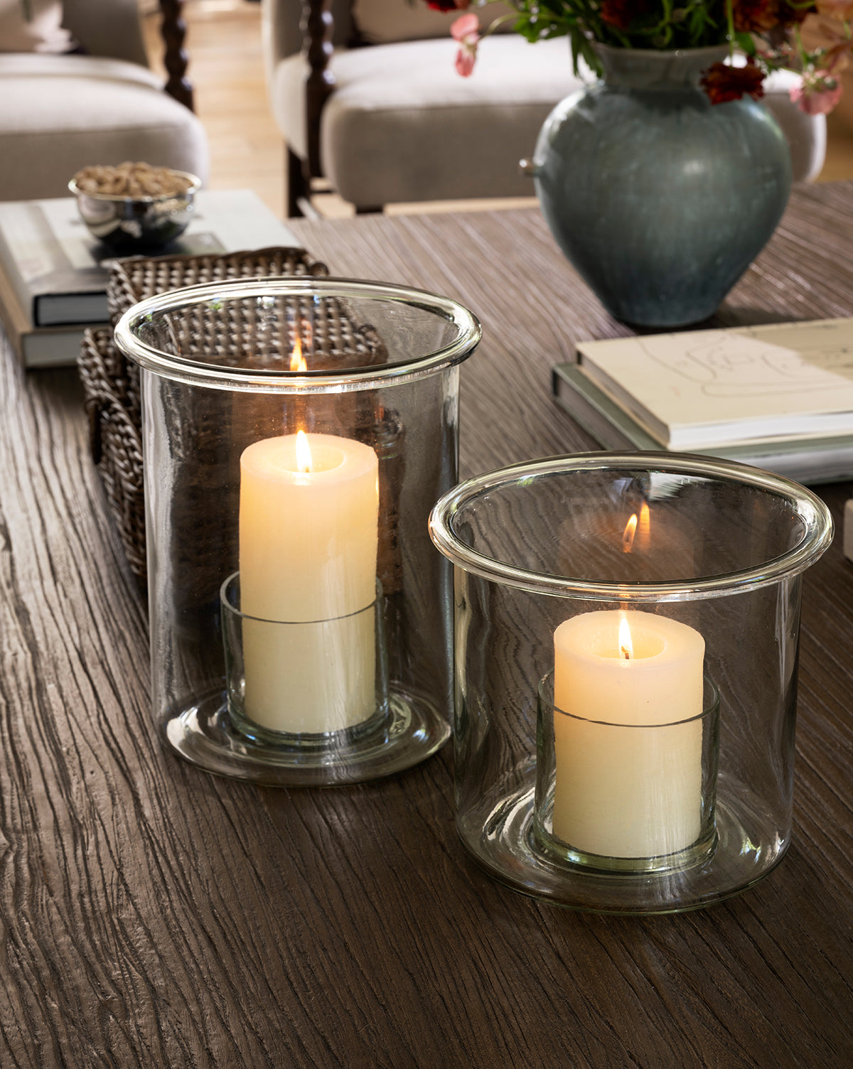 Two lit pillar candles in Cyrus Hurricane holders sit on a wooden table. Nearby are a ceramic vase with flowers, stacked books, a woven basket, and snacks, creating a cozy, inviting home decor atmosphere by McGee & Co.