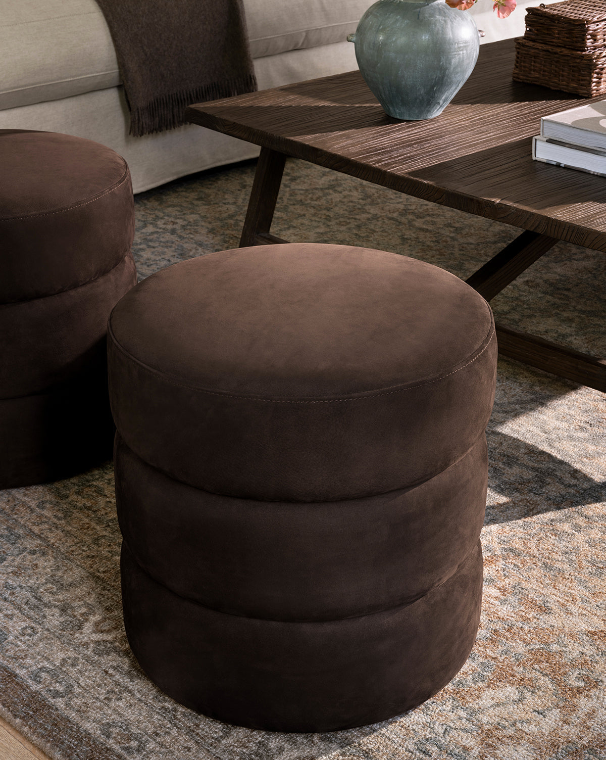 The Channel Ottoman, featuring a round shape, dark brown suede, and quilted design, rests on a patterned rug next to a wooden coffee table in a cozy midcentury living room, by McGee & Co.