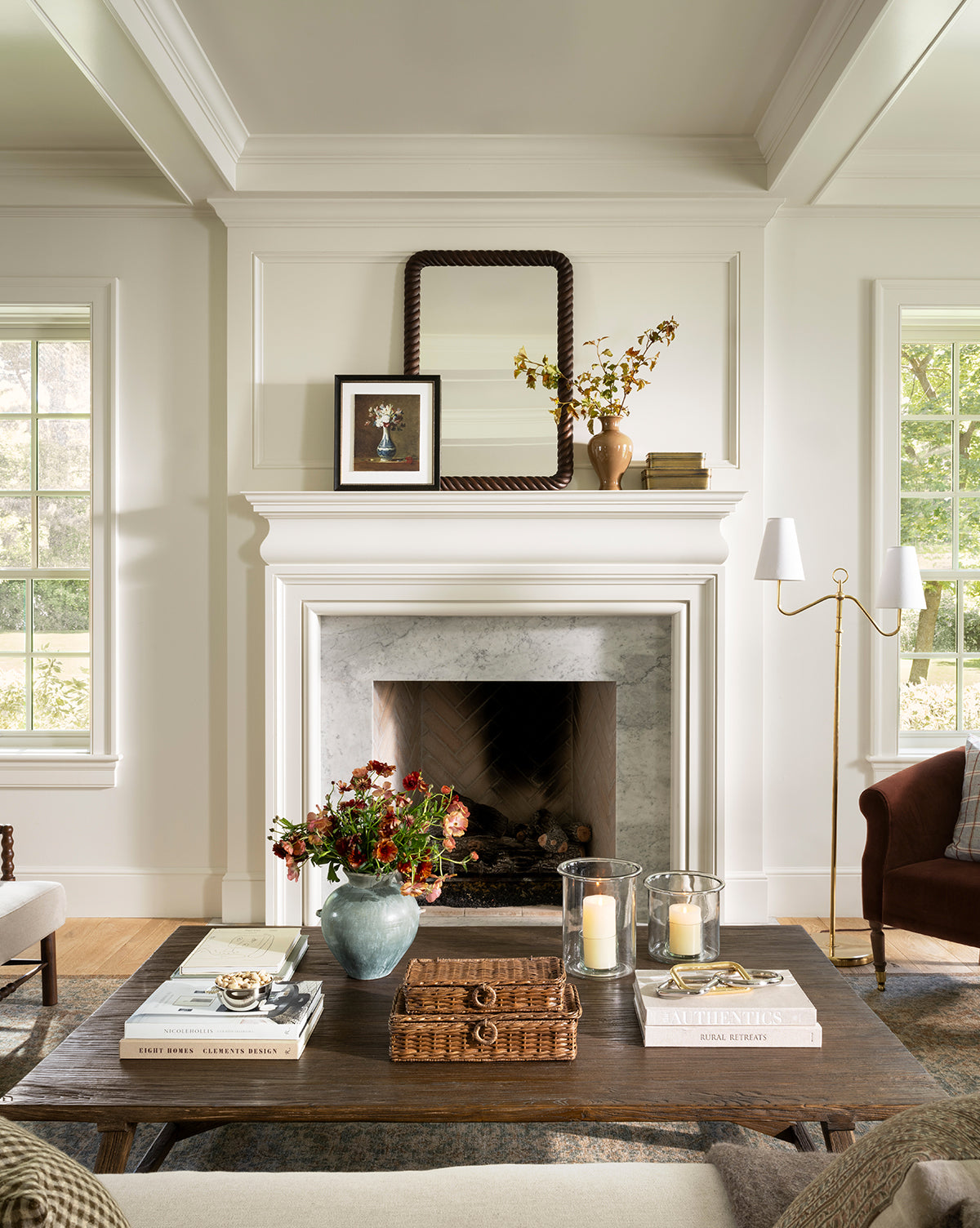 A cozy, elegant living room with a white fireplace, a mirror and vintage-style wall art above the mantel, a wooden coffee table displaying an Antique Flower Vase, and large windows letting in natural light by McGee & Co.