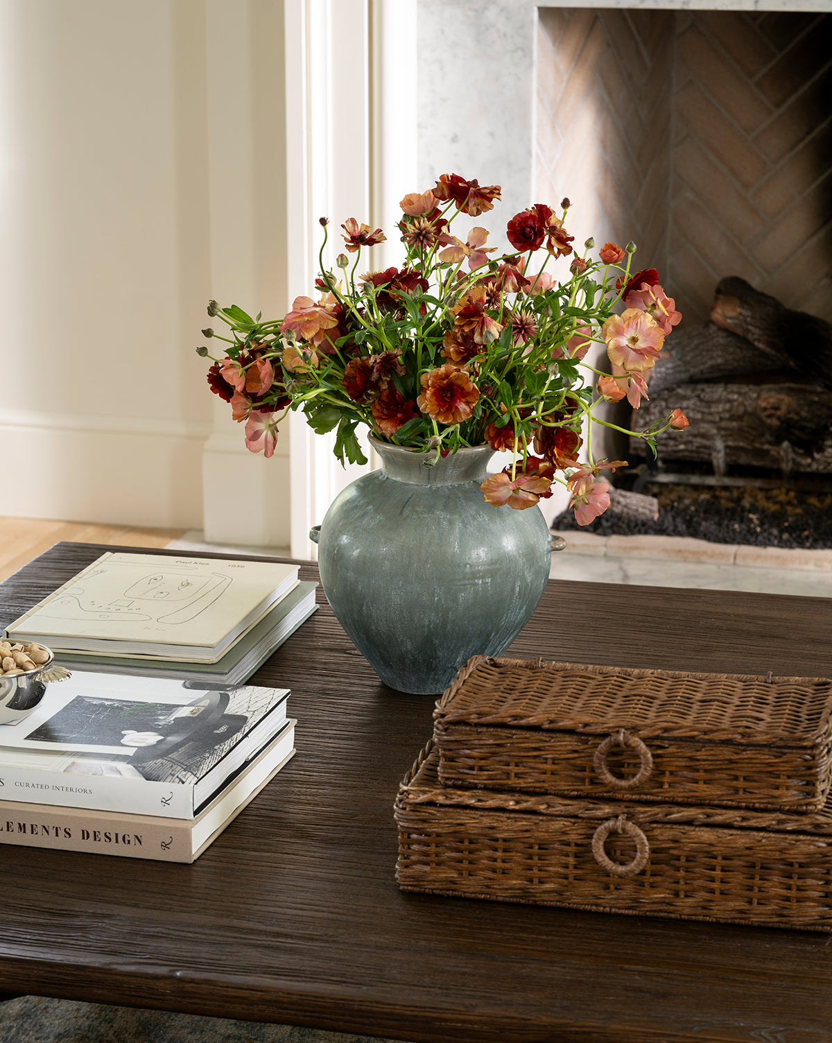 A Beatrix Vase brimming with red and pink blooms sits on a dark wood coffee table next to wicker boxes, books, and papers, with a fireplace behind it—showcasing the vintage-inspired handcrafted artistry of the Beatrix Vase, by McGee & Co.