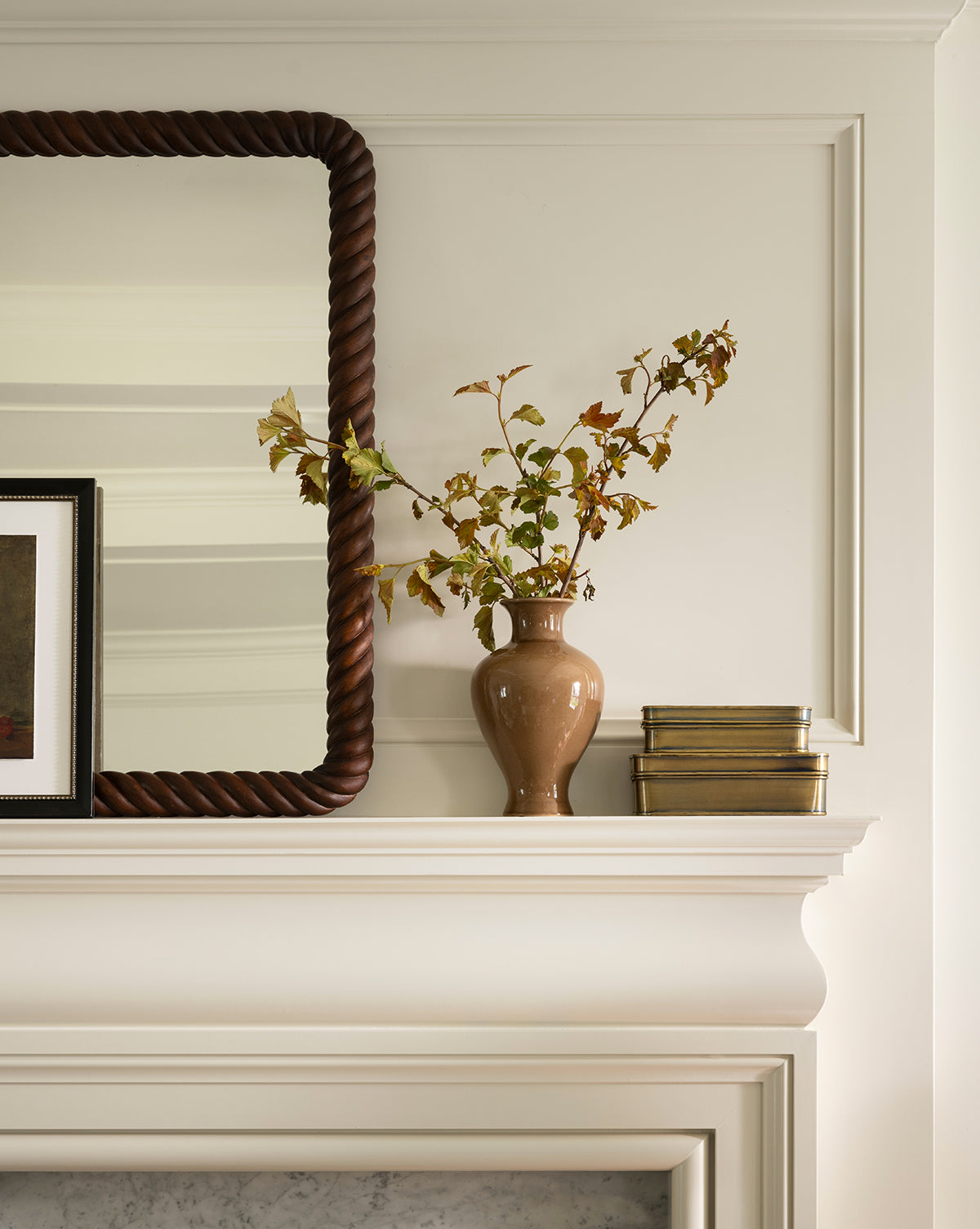 The Natalia Vase, filled with leafy branches, sits on a white mantel next to two gold boxes, with a large mirror in a twisted dark frame and a framed picture beside it, by McGee & Co.