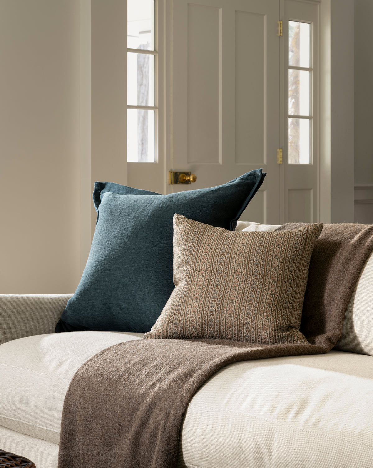 A cozy beige sofa with a dark blue pillow, the Wakefield Pillow Cover in a patterned brown design, and a brown throw blanket, set in a softly lit room with white paneled doors and large windows in the background, by McGee & Co.