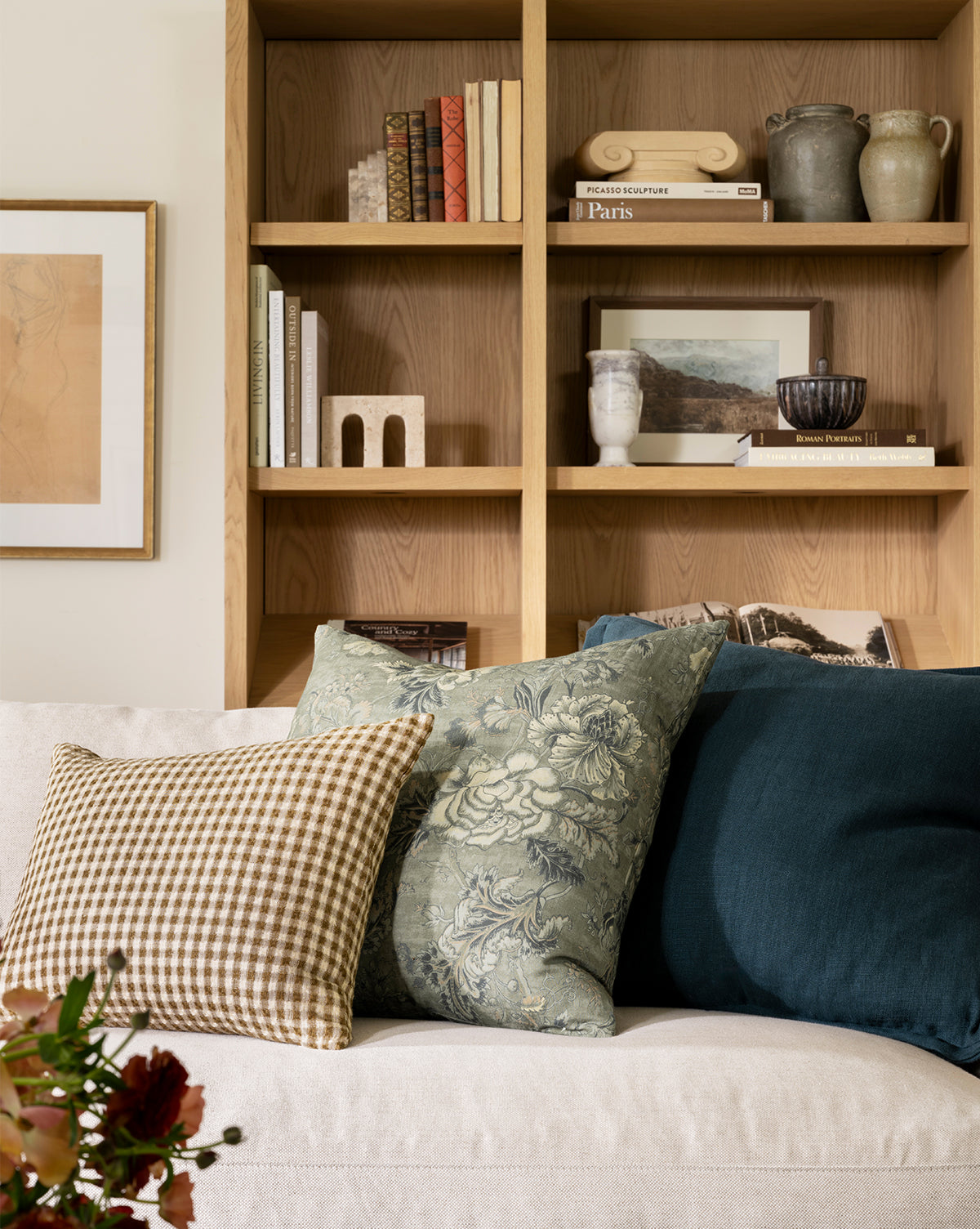 A beige sofa with patterned and solid throw pillows, including the Liam Double Flange Pillow Cover, sits in front of a wooden bookshelf filled with books and decor. Framed artwork hangs nearby, by McGee & Co.