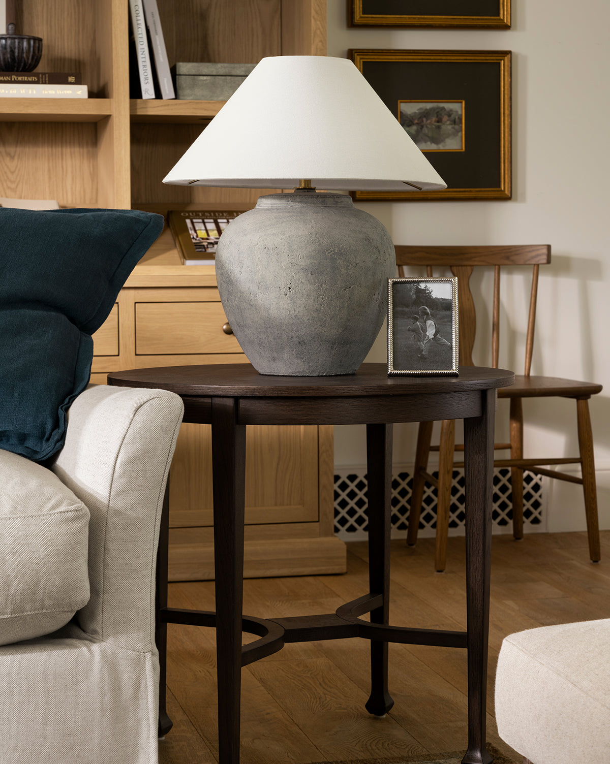 A round dark wood side table with the Montague Table Lamp, a framed black-and-white photo, and books stands next to a light sofa with a teal cushion in a cozy living room, by McGee & Co.