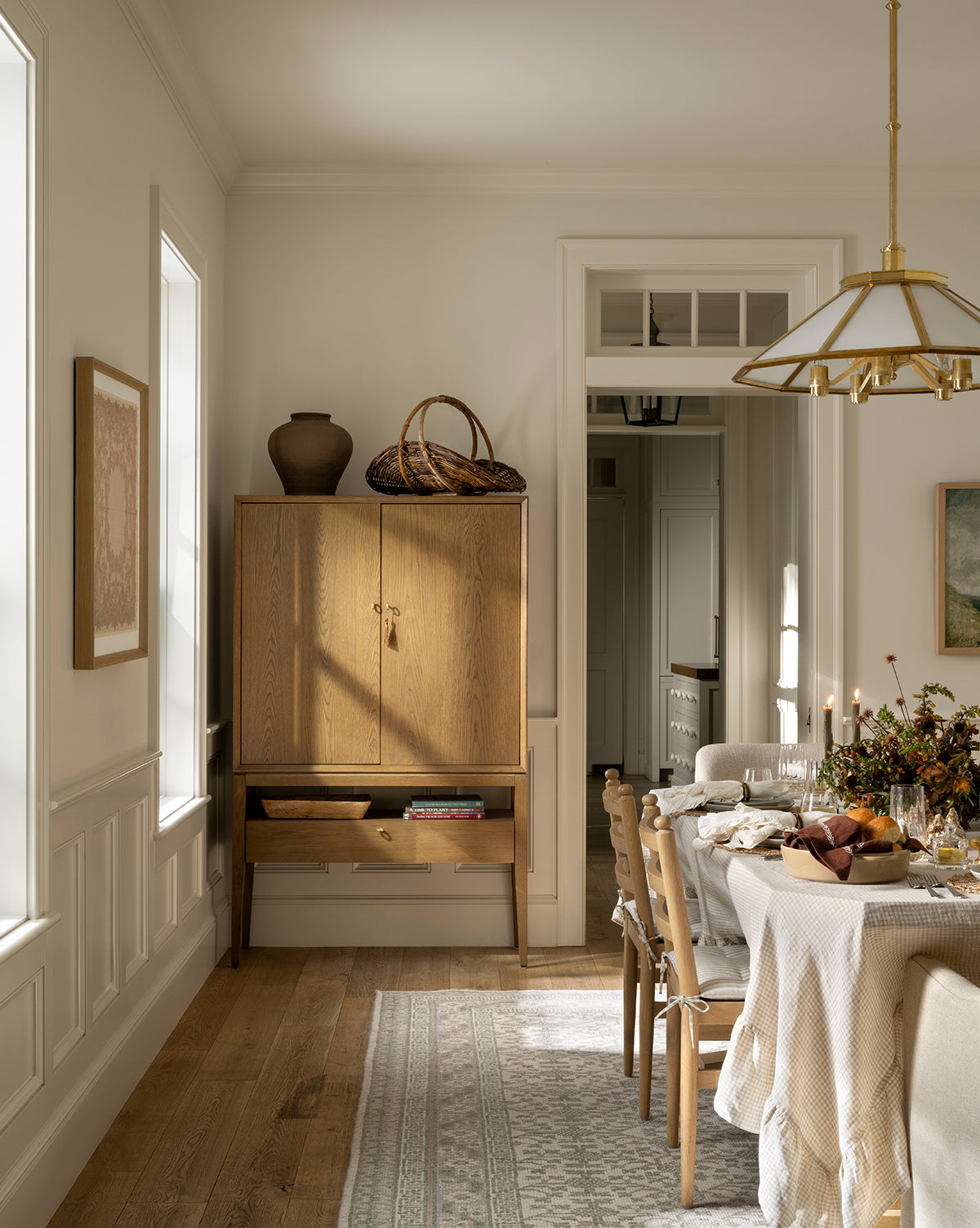 A bright dining room features a wooden table set for a meal with chairs, while the Orsino Hand-Knotted Rug adds warmth beneath. A light wood cabinet and large windows complete the cozy, neutral space by McGee & Co.