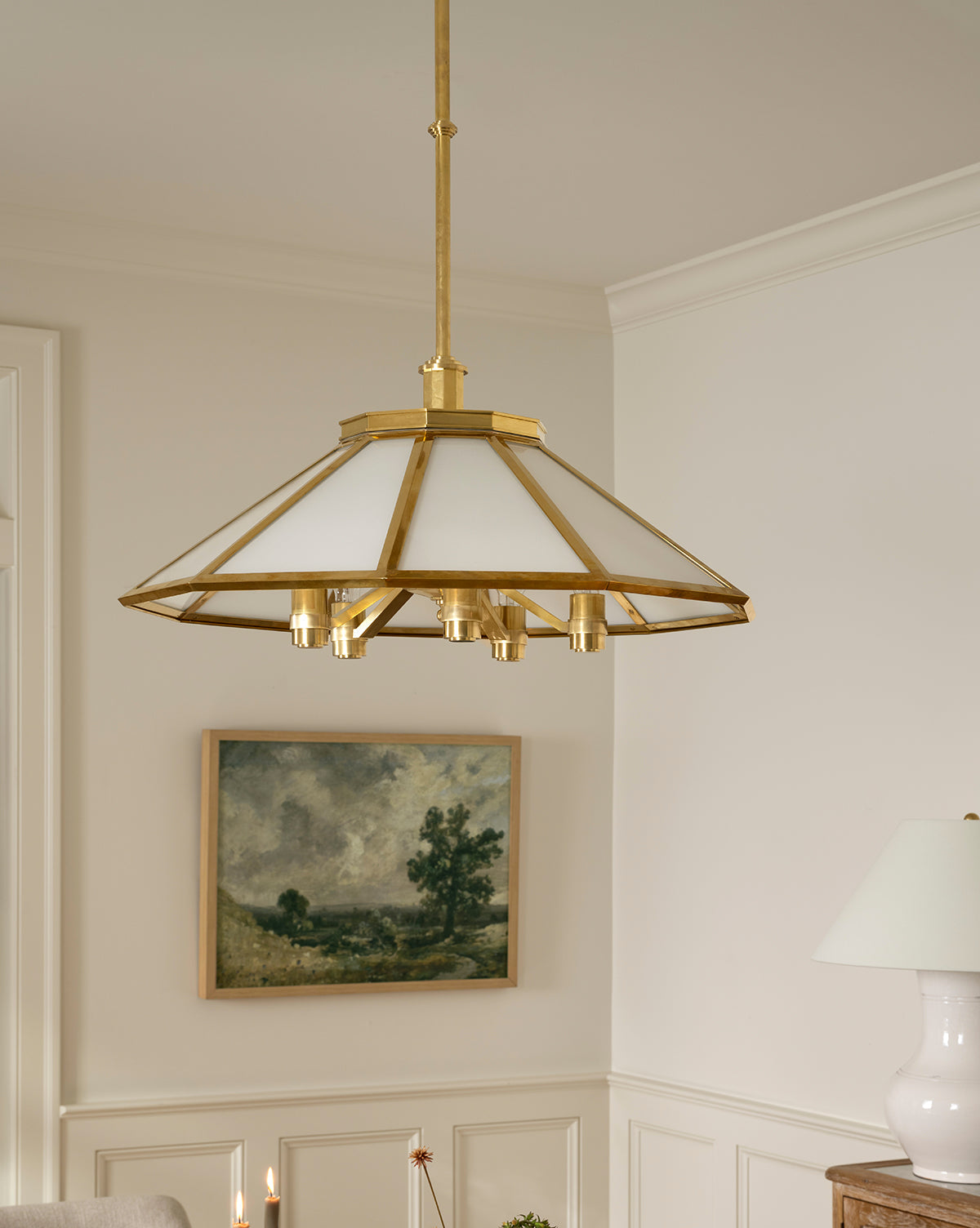 The Knightley Chandelier with its geometric antique brass design hangs above a neutral-toned room, while a framed landscape artwork and a white table lamp add contrast to the light-colored walls, by McGee & Co.
