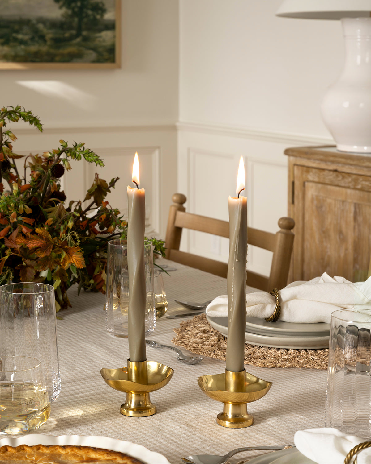 Two lit Errol Wax Tapers (Set of 6) in brass holders sit on a dining table with glassware, woven placemat, white napkins, and a fall floral arrangement. A sideboard and framed art are visible in the background, by McGee & Co.