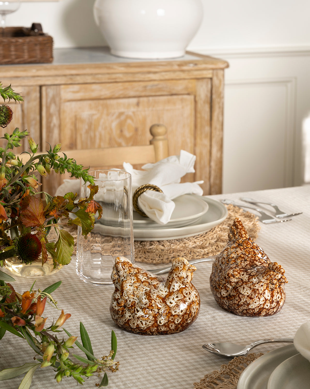 A dining table set with neutral plates, glasses, and napkins displays artisanal charm with a Stoneware Hens (Set of 2) centerpiece amid autumn foliage; a wooden sideboard appears in the background, by McGee & Co.