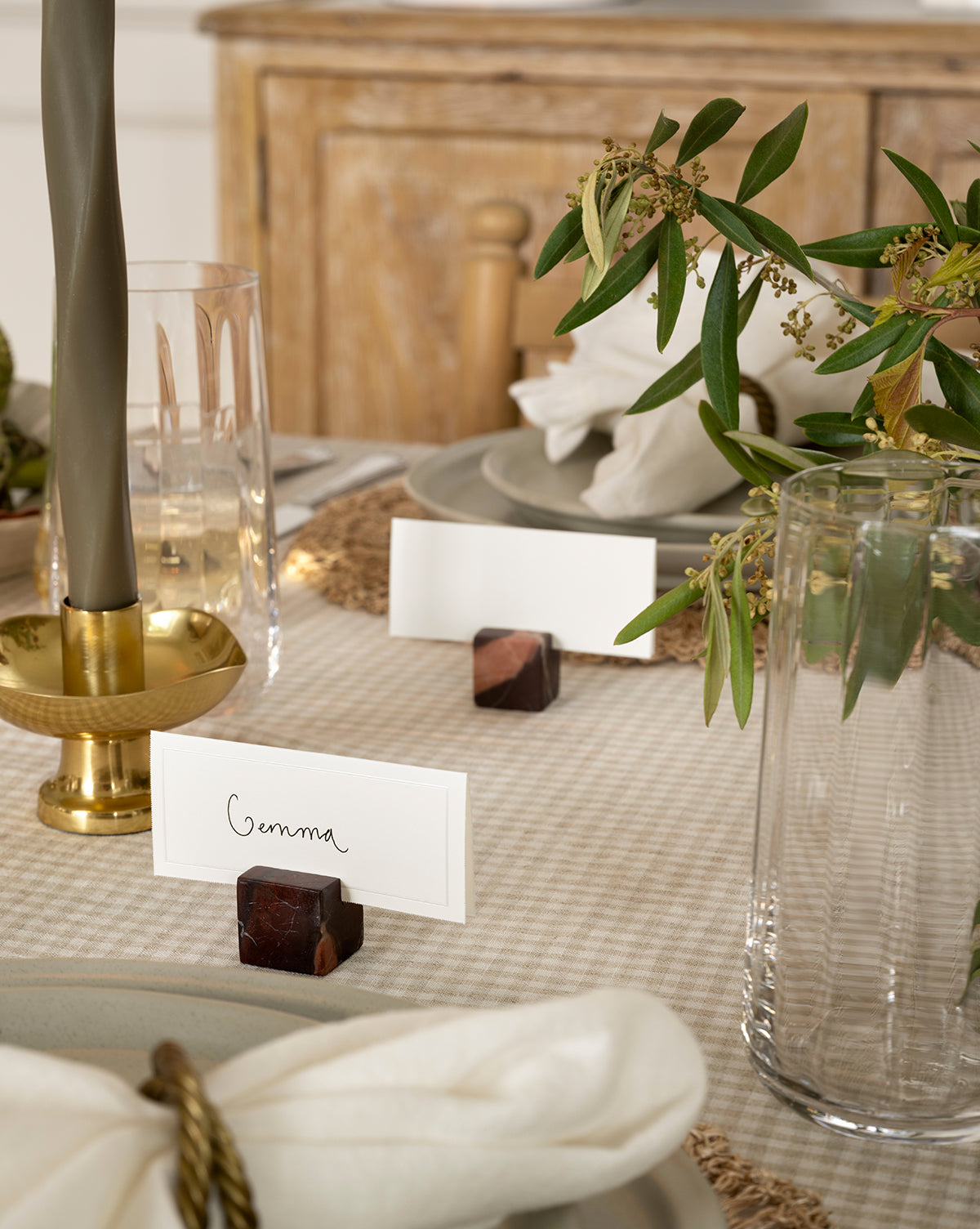 A close-up of an elegant harvest tablescape featuring the Auburn Marble Place Card Holders (Set of 4) with a name card, gold candlestick, clear glassware, white napkin, and green foliage on a beige checkered tablecloth—perfect for holiday gatherings, by McGee & Co.