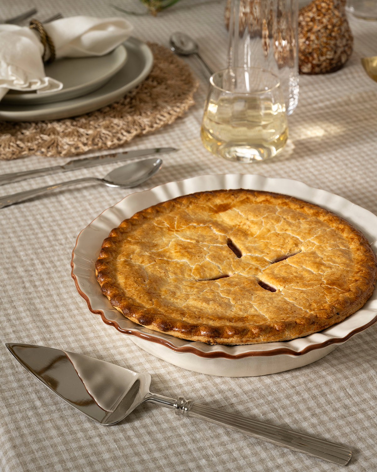 A golden-brown pie rests in a white ceramic dish on a checked tablecloth, accompanied by the McNully Pie Server, silver cutlery, a glass of white wine, and a neatly set plate with a folded napkin by McGee & Co.