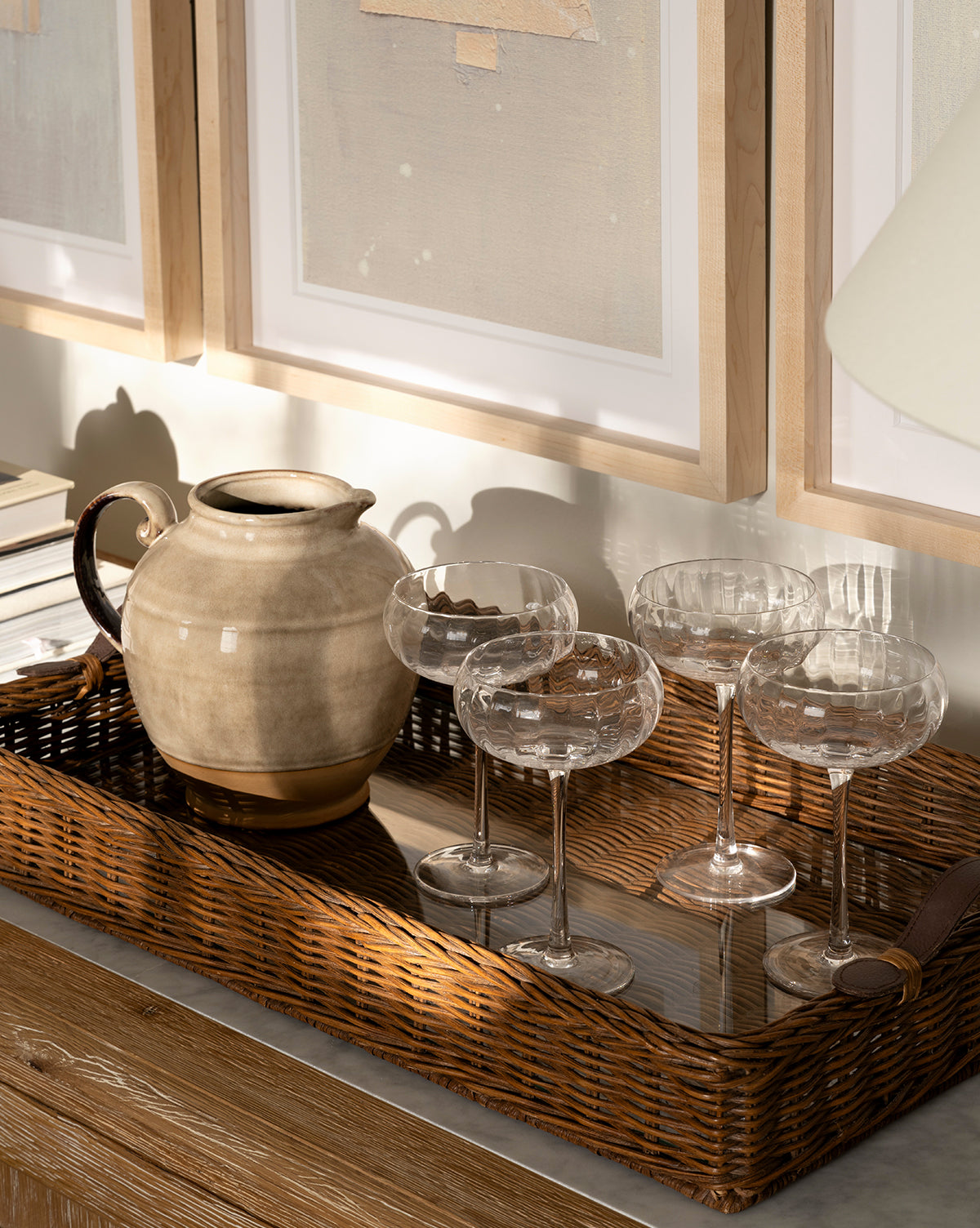 A wicker tray on a wooden surface displays the Brown Stoneware Pitcher and four empty coupe glasses. Framed abstract art and soft sunlight in the background create gentle shadows, offering perfect kitchen styling inspiration, by McGee & Co.