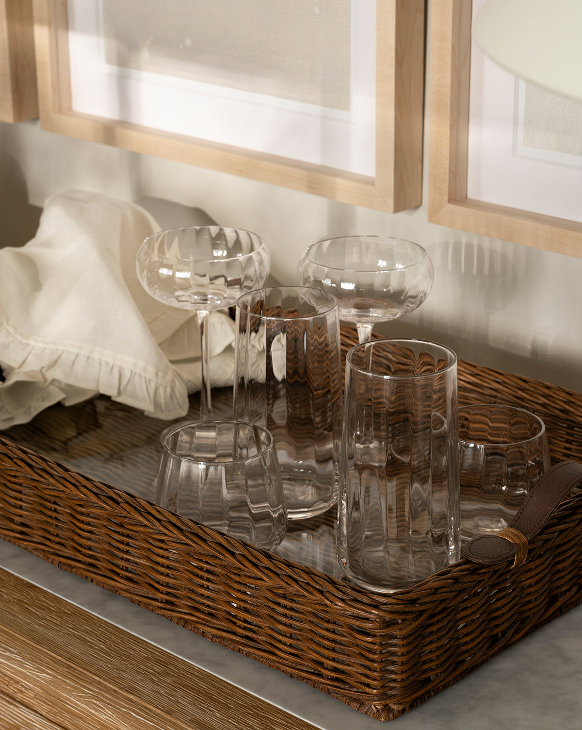 A wicker tray on a marble surface holds the Langford Tumblers (Set of 4) in assorted shapes and sizes, a folded cream-colored napkin, and is set against a wall adorned with framed art, by McGee & Co.