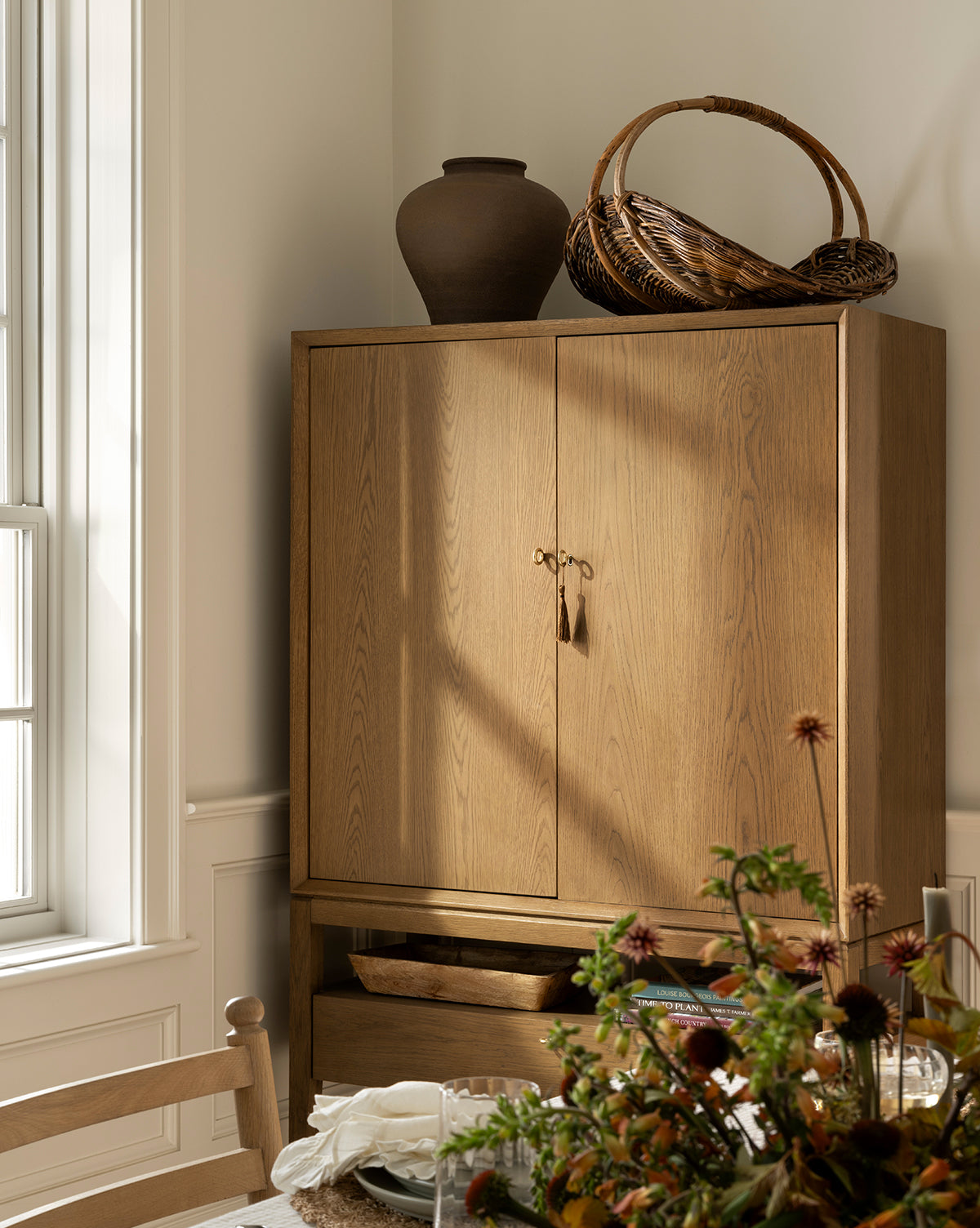 A wooden cabinet with closed doors stands in a sunlit room, topped with a matte brown Reading Vase and a woven basket. A dining table with flowers, napkins, and glassware is partially visible in the foreground, creating inviting home decor by McGee & Co.