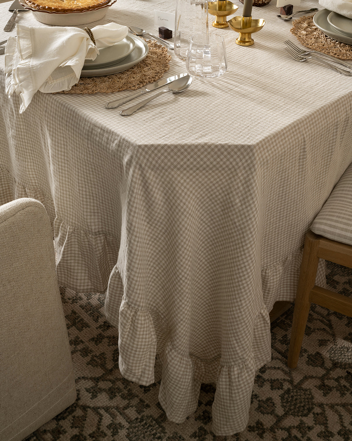 A dining table set with plates and brass candlesticks is covered by the Catania Linen Ruffle Tablecloth in beige gingham, its ruffled edges showing, alongside a glimpse of a chair and patterned rug—creating a charming atmosphere, by McGee & Co.