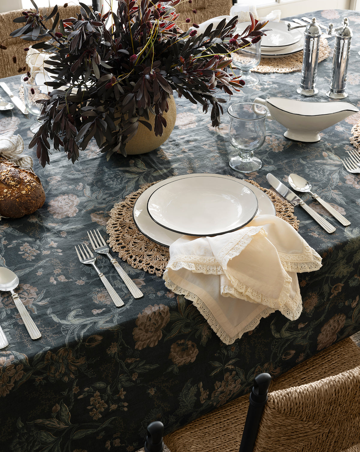 A dining table set with a floral tablecloth, white plates on Hermione Placemat, silver cutlery, folded lace-edged napkins, clear glasses, bread, and a vase of dark leafy branches as the centerpiece, by McGee & Co.