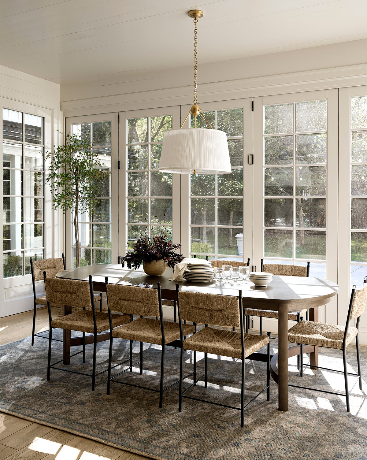 A bright dining room showcases the Gardiner Dining Table with eight woven chairs on a patterned rug, illuminated by large windows. A potted plant and chandelier complete the space, by McGee & Co.