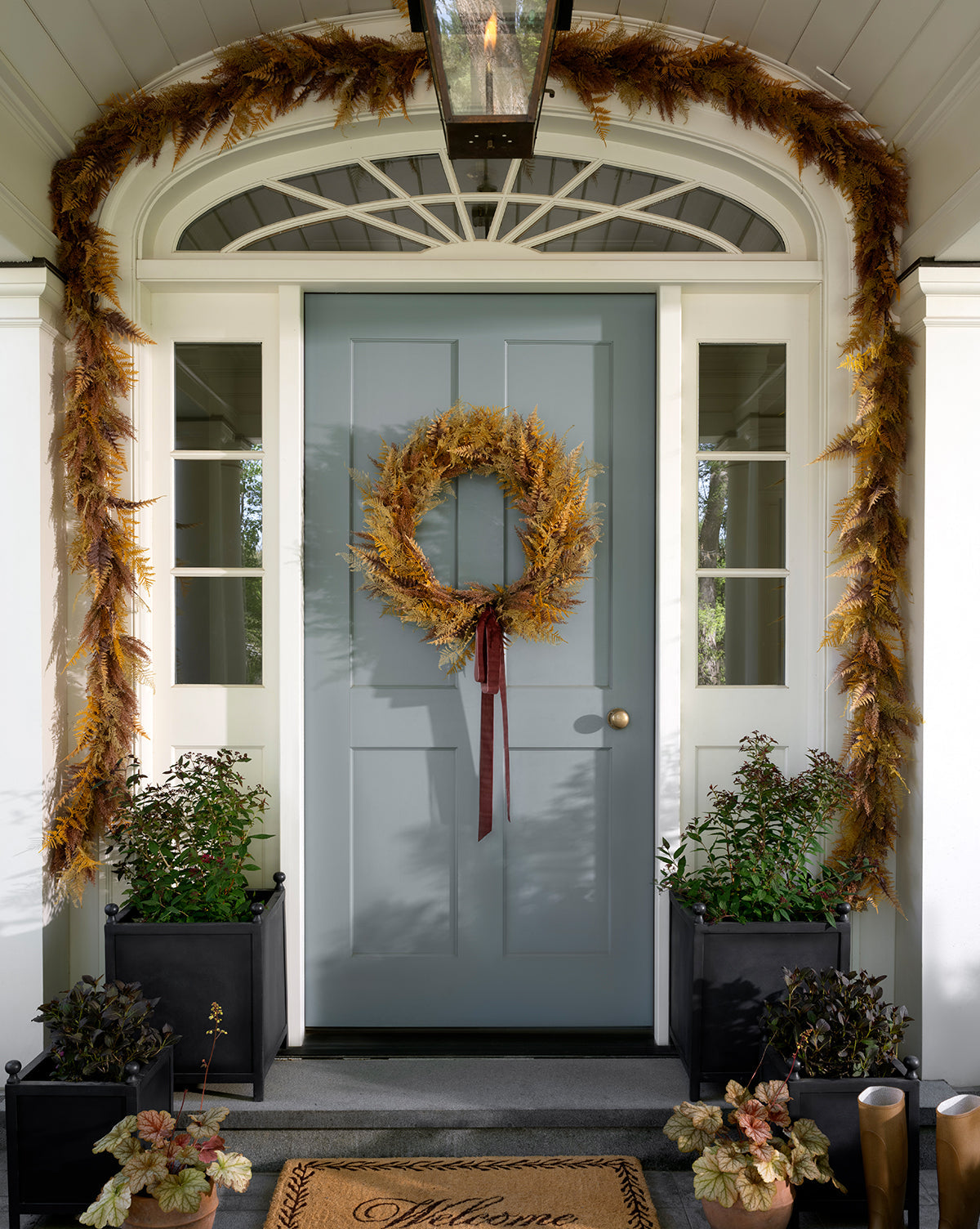 A blue front door displays the 30 Faux Golden Fern Wreath with coordinating garland, creating a stunning entryway. Black planters with leafy greens flank the Welcome doormat, all framed by elegant white columns, by McGee & Co.