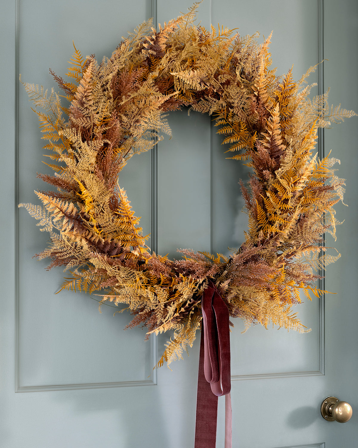 The 30 Faux Golden Fern Wreath features dried golden-brown ferns and a velvet burgundy ribbon, hanging on a light blue door with a brass round doorknob—an elegant fall wreath that adds timeless entryway charm, by McGee & Co.