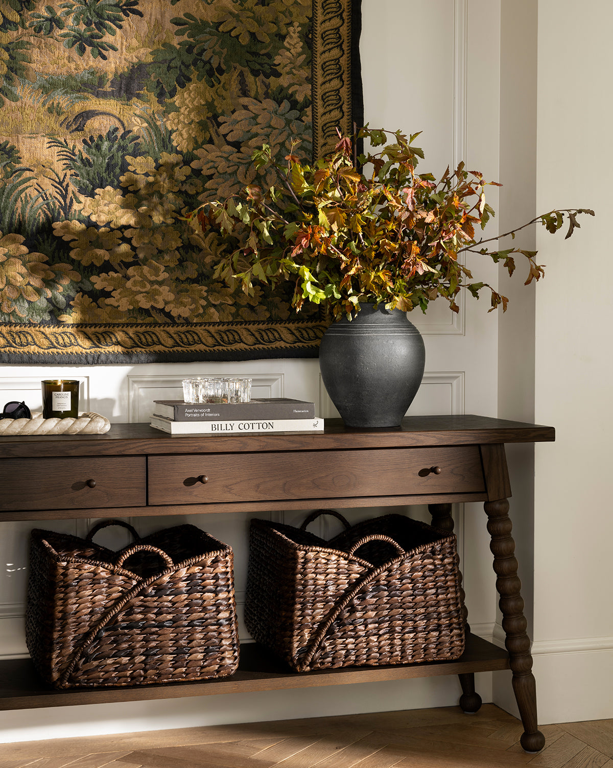 A dark wooden console table with a black vase, books, and candles sits below a tapestry. Two Quimby Baskets (Set of 2) are placed on the lower shelf against a white wall, by McGee & Co.