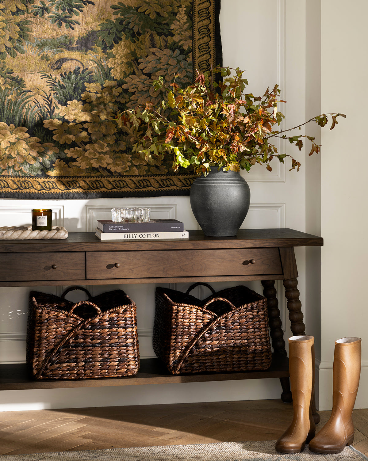 A wooden console table displays the Ethel Vase filled with autumn branches, alongside books and glassware. Woven baskets sit below, tall tan boots rest in front, and a tapestry provides rustic decor on the wall behind, by McGee & Co.