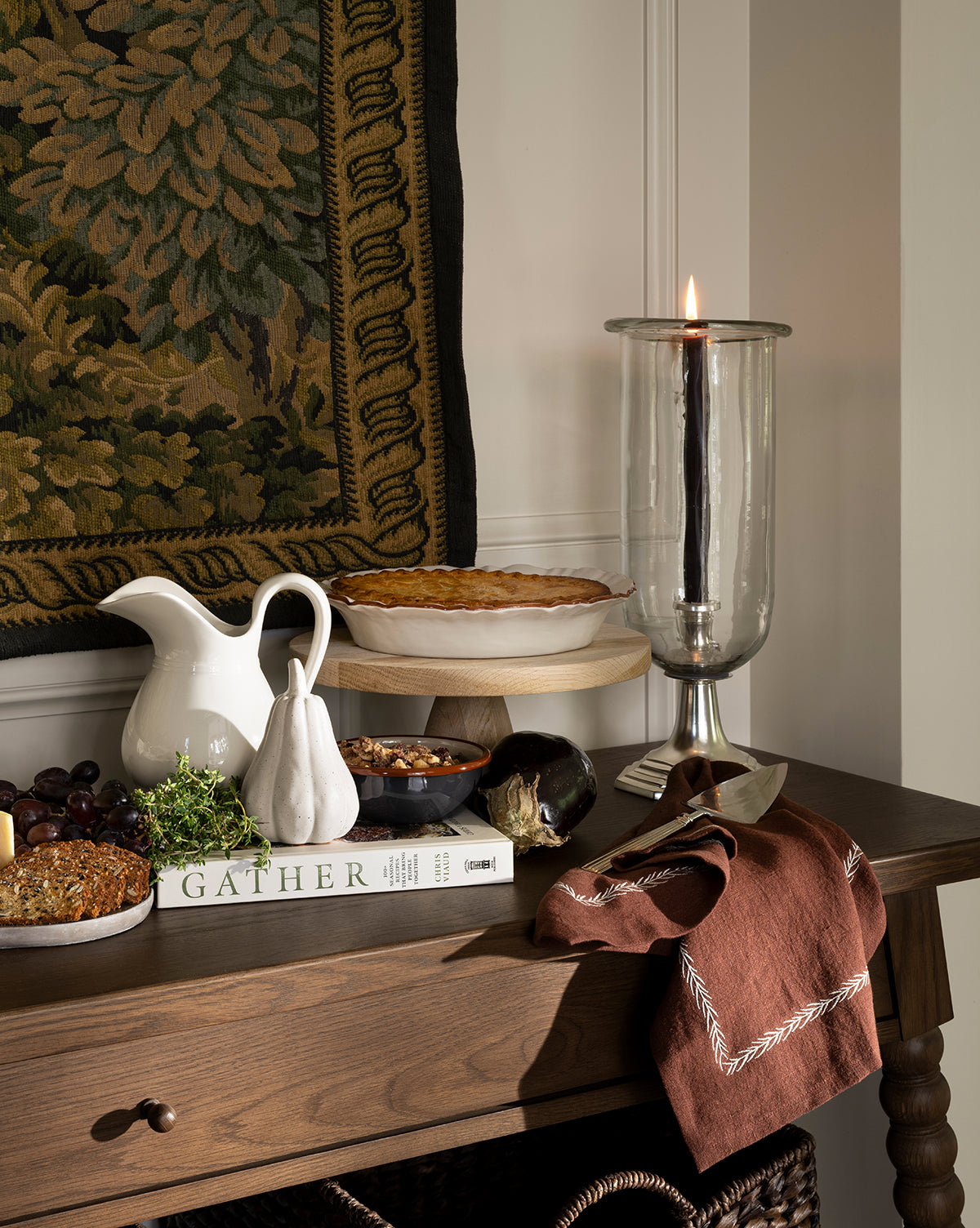 A wooden table displays a pie, fruit, nuts, a white pitcher, ceramic pear, brown cloth napkin, Errol Wax Tapers (Set of 6) in glass holders, and a book titled GATHER against a tapestry and white paneled wall by McGee & Co.