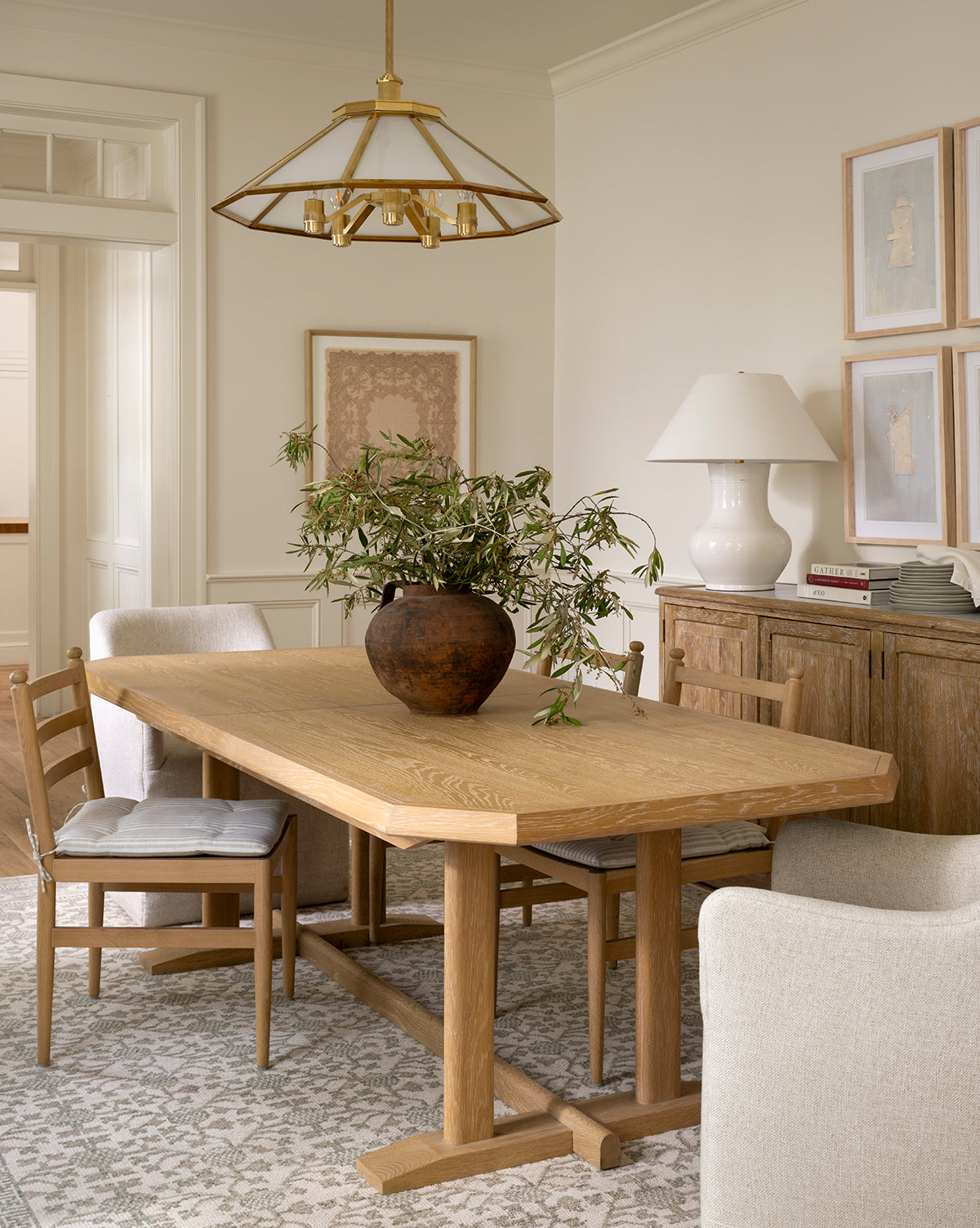 The European Flare dining room features a wooden table, cushioned chairs, a ceramic vase with greenery, sideboard, white lamp, vintage-style wall art in a woodgrain frame, neutral tones, and a brass chandelier for classic charm, by McGee & Co.