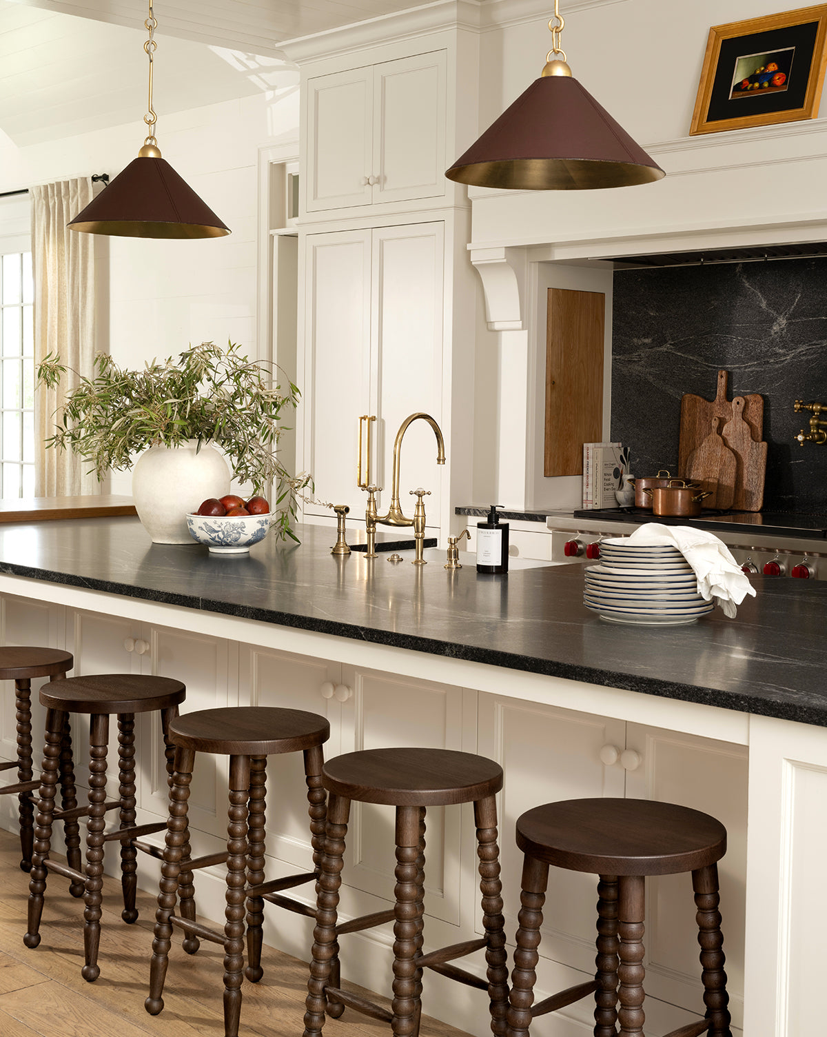 A modern kitchen with a black countertop island, five dark wooden stools, gold fixtures, pendant lights, curated decor including stacked plates, a white vase with greenery, a bowl of apples, and Autumns Bounty I by Pascal Giroud art, by McGee & Co.