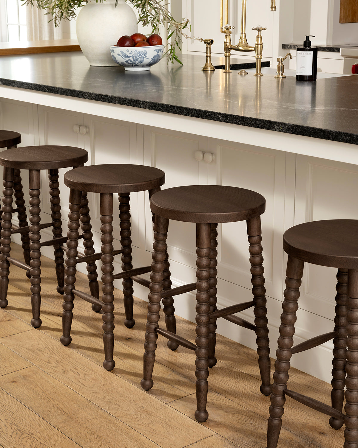 Five Heathcliff Stools with dark wooden, bobbin-style turned legs are lined up along a kitchen island topped with a black counter. A bowl of fruit, gold faucet, and soap dispenser sit on the counter; the floor is light wood, by McGee & Co.